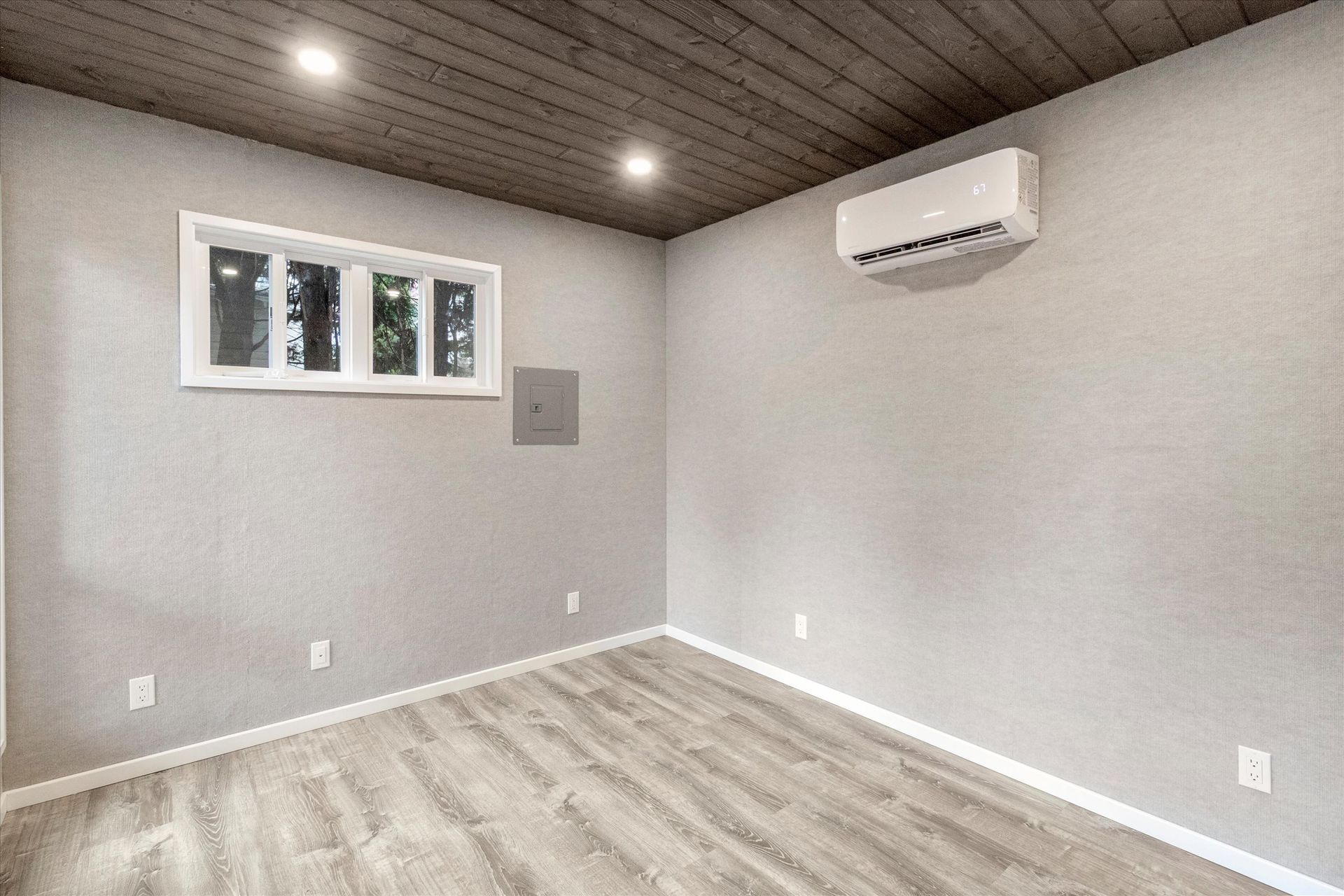 A minimalist room with light grey walls, wood-look flooring, a window, and a wall-mounted AC unit under a wood-plank ceiling.