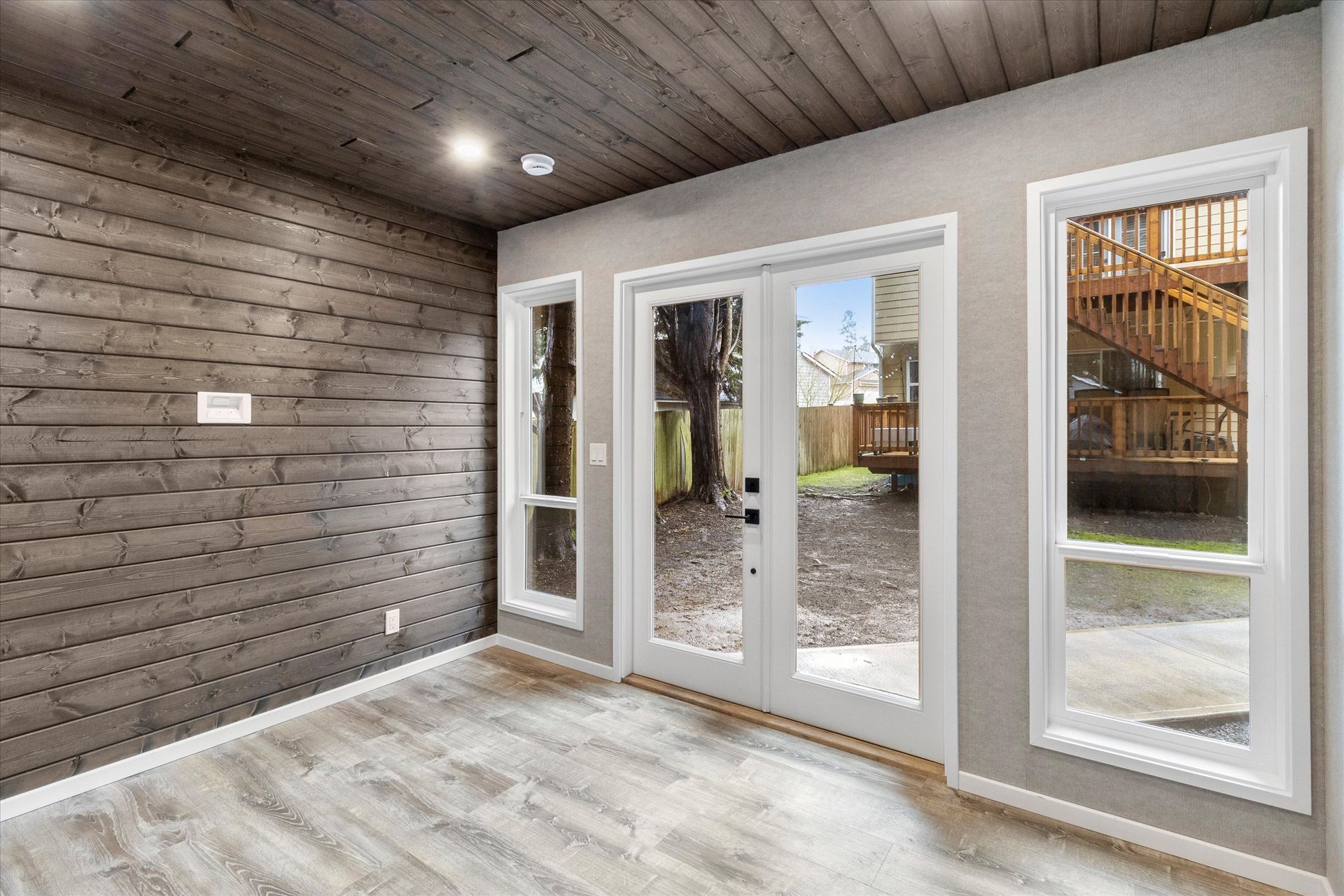 A modern room with grey horizontal wood-paneled walls and ceiling, light wood floors, glass French doors, and windows.