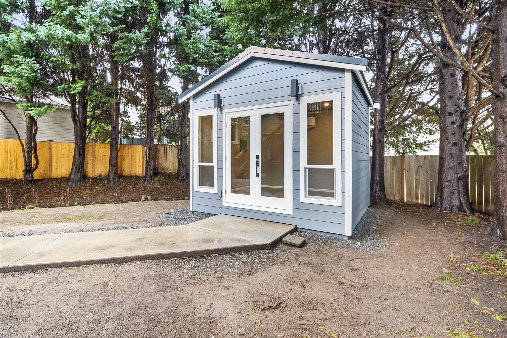 A light blue modern backyard shed with double glass doors and vertical side windows, set on a gravel path near trees.