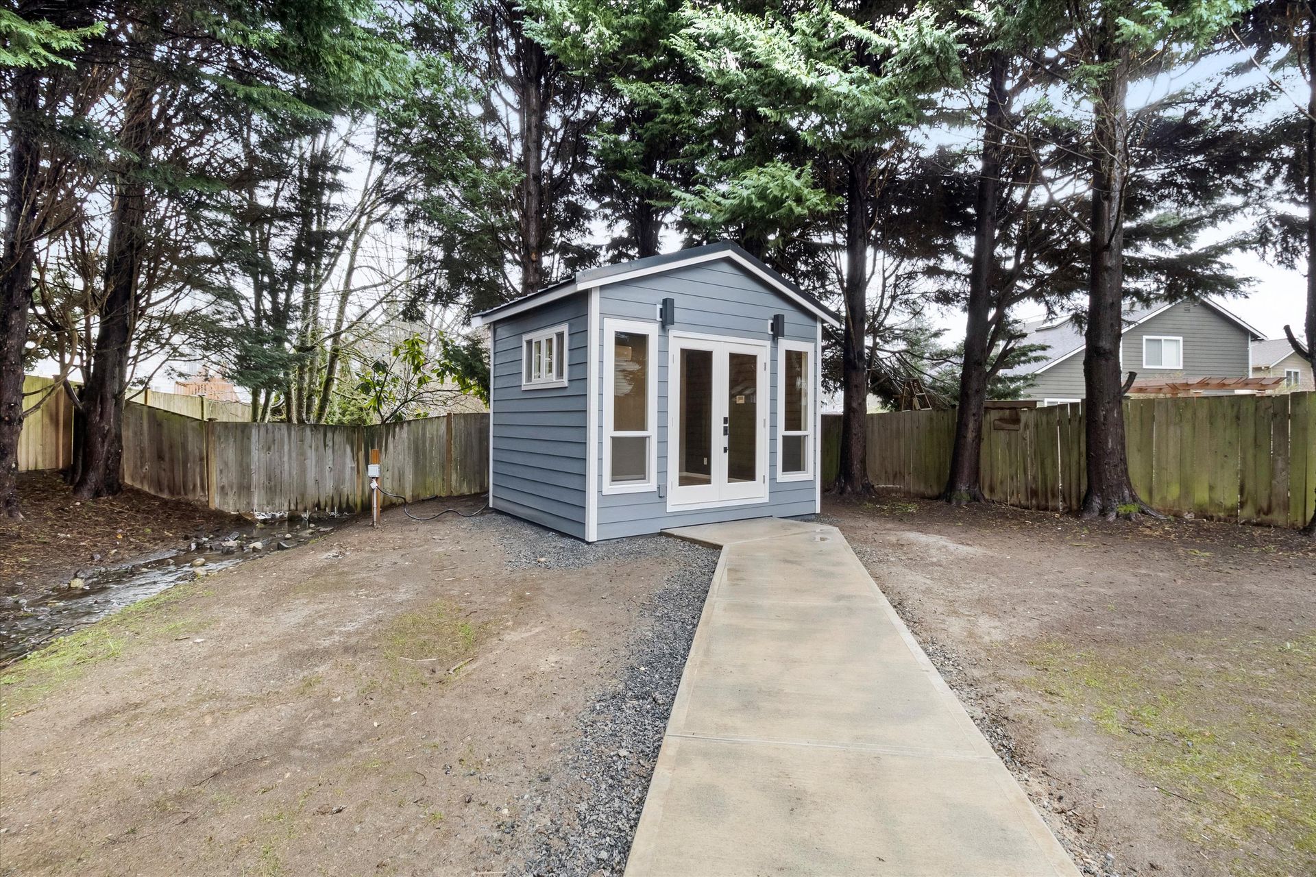 A grey garden shed with French doors and windows sits in a yard with a concrete path, surrounded by a wooden fence and trees.