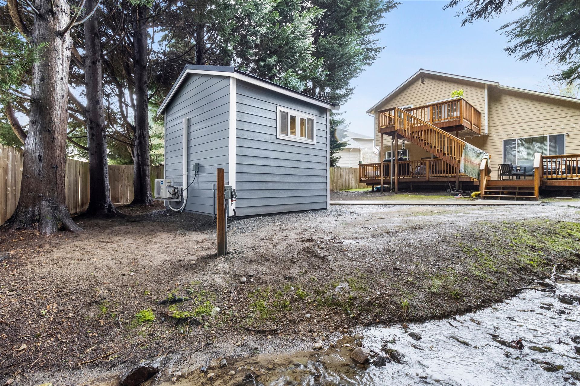 A grey shed stands in a wooded backyard near a beige house with a wooden deck and exterior staircase.
