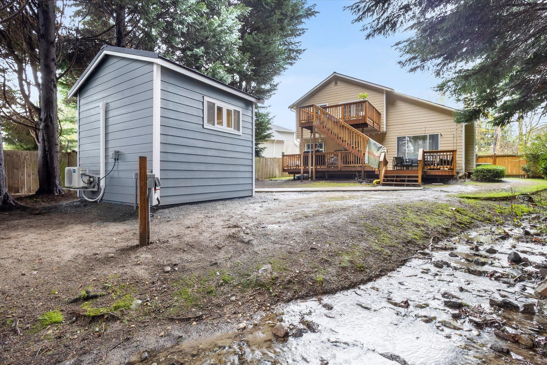 A gray shed in a yard, with a wooden deck and staircase of a house visible in the background.