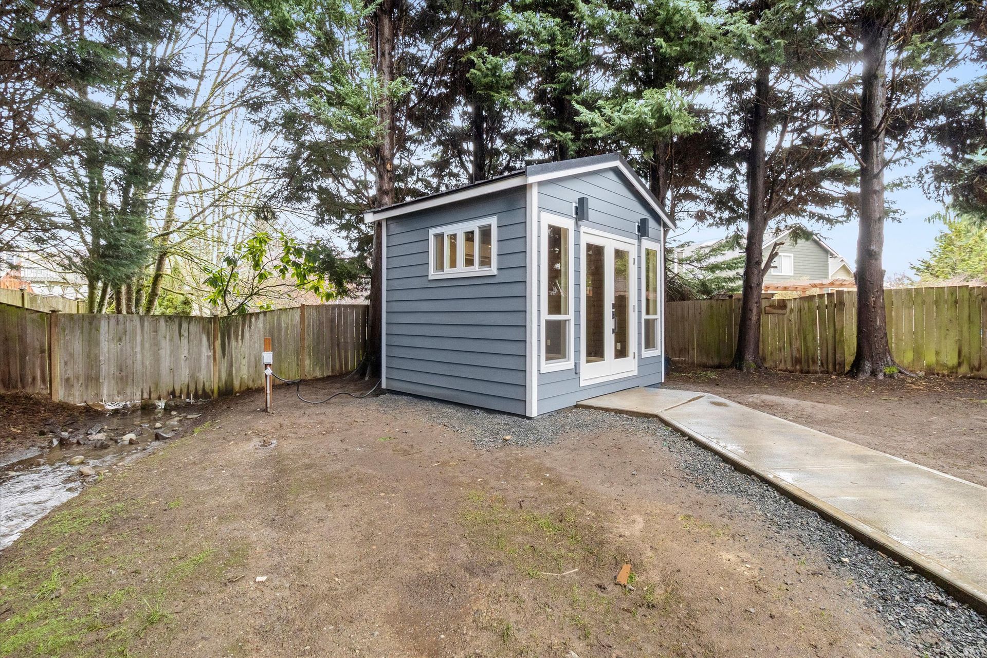 A small, grey backyard shed with glass doors and windows, situated on a patch of dirt and grass next to a concrete path.