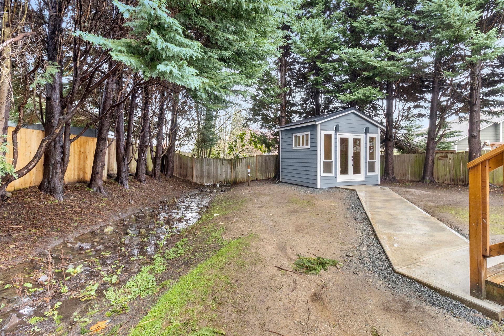 A gray shed with a glass door sits at the end of a paved path in a backyard with tall trees and a muddy, wet area.