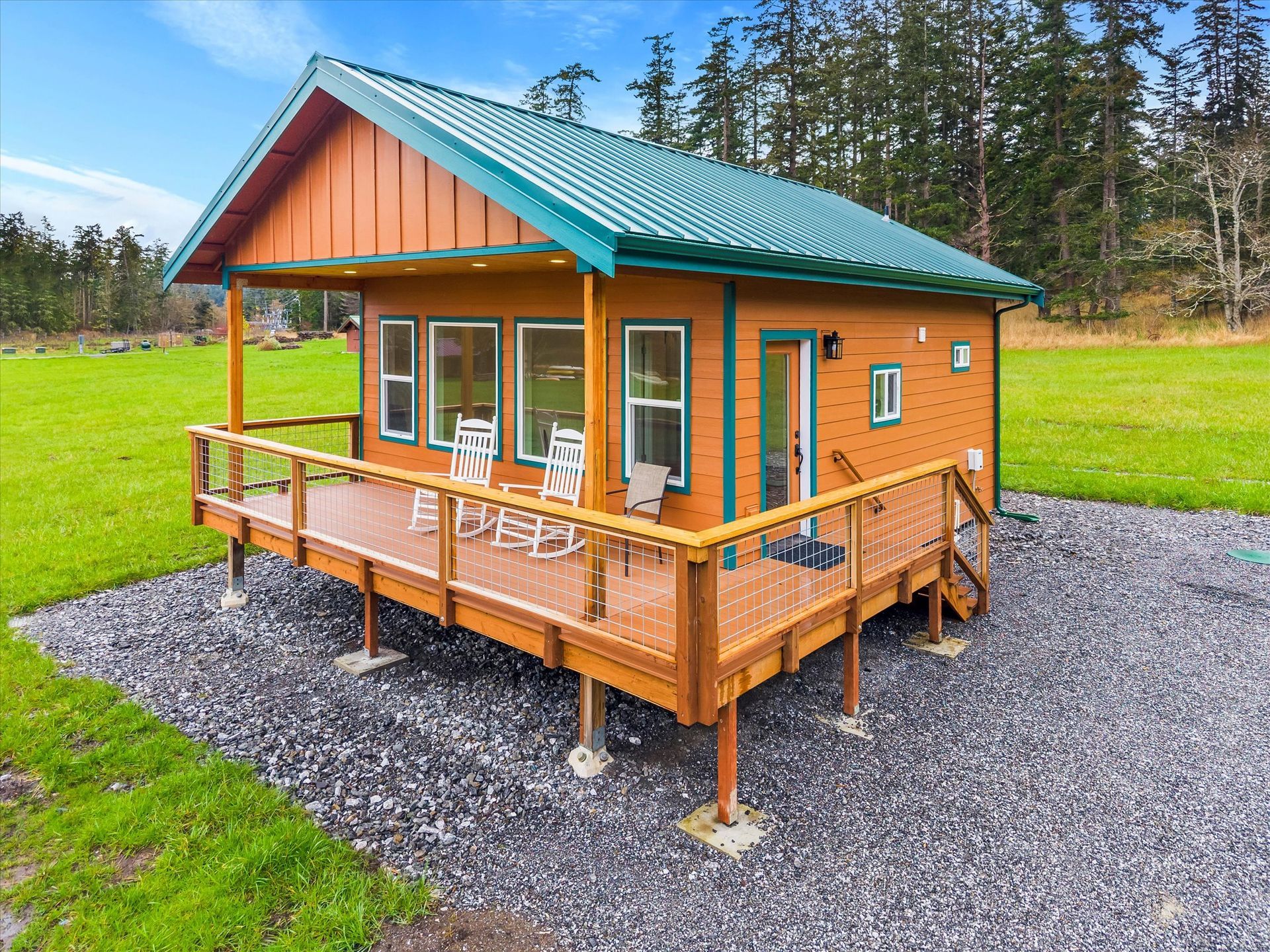 Small wood-sided cabin with a green metal roof, wrap-around deck, and two rocking chairs in a grassy field.