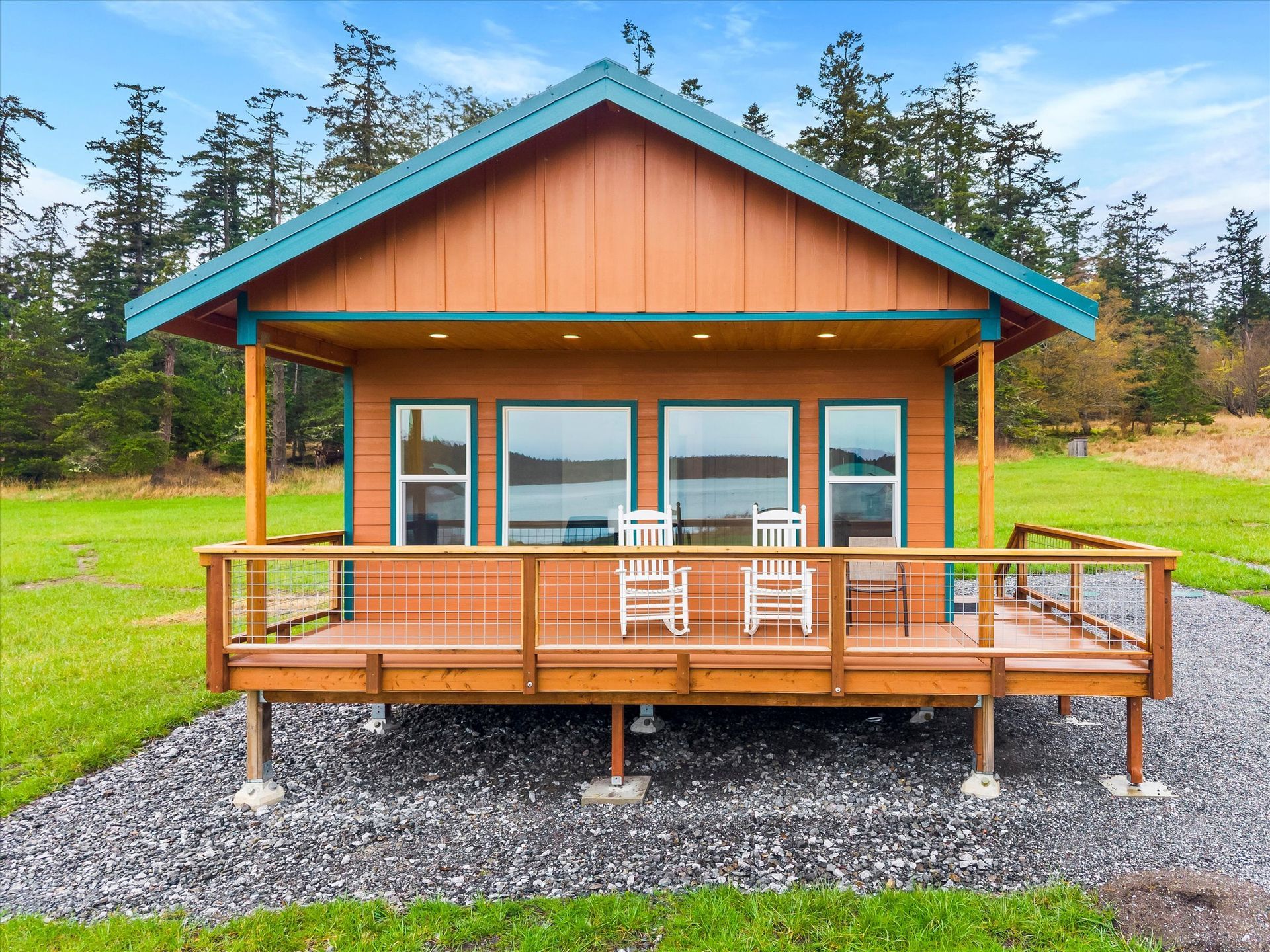 A wooden cabin with a teal roof and a front deck sits on a gravel patch overlooking a body of water amidst green fields.