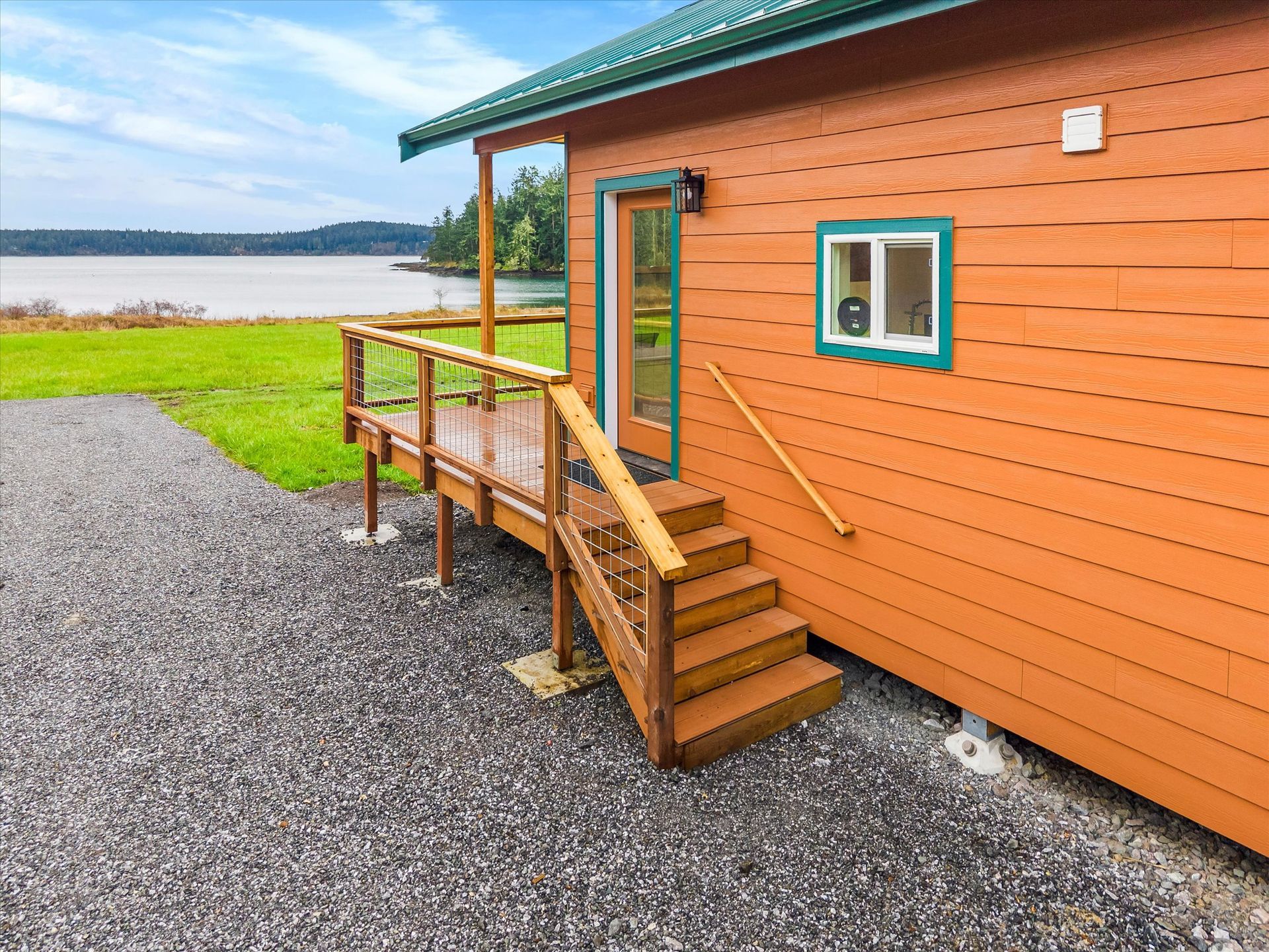 An orange cabin with a green-trimmed door and window sits on a gravel lot overlooking a calm lake and grassy shore.