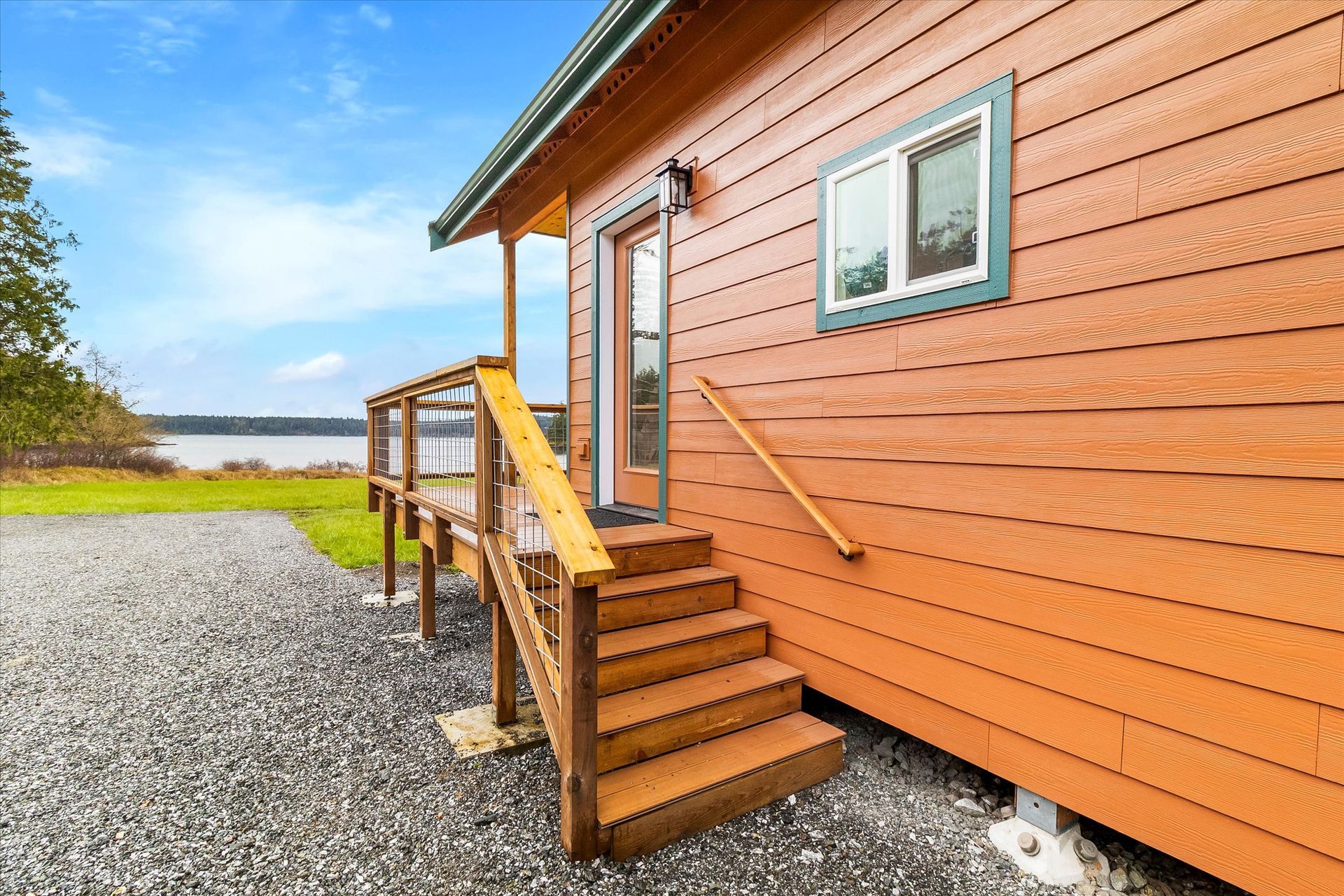 A wooden cabin with orange horizontal siding, stairs leading to a deck, and a view of a body of water under a blue sky.