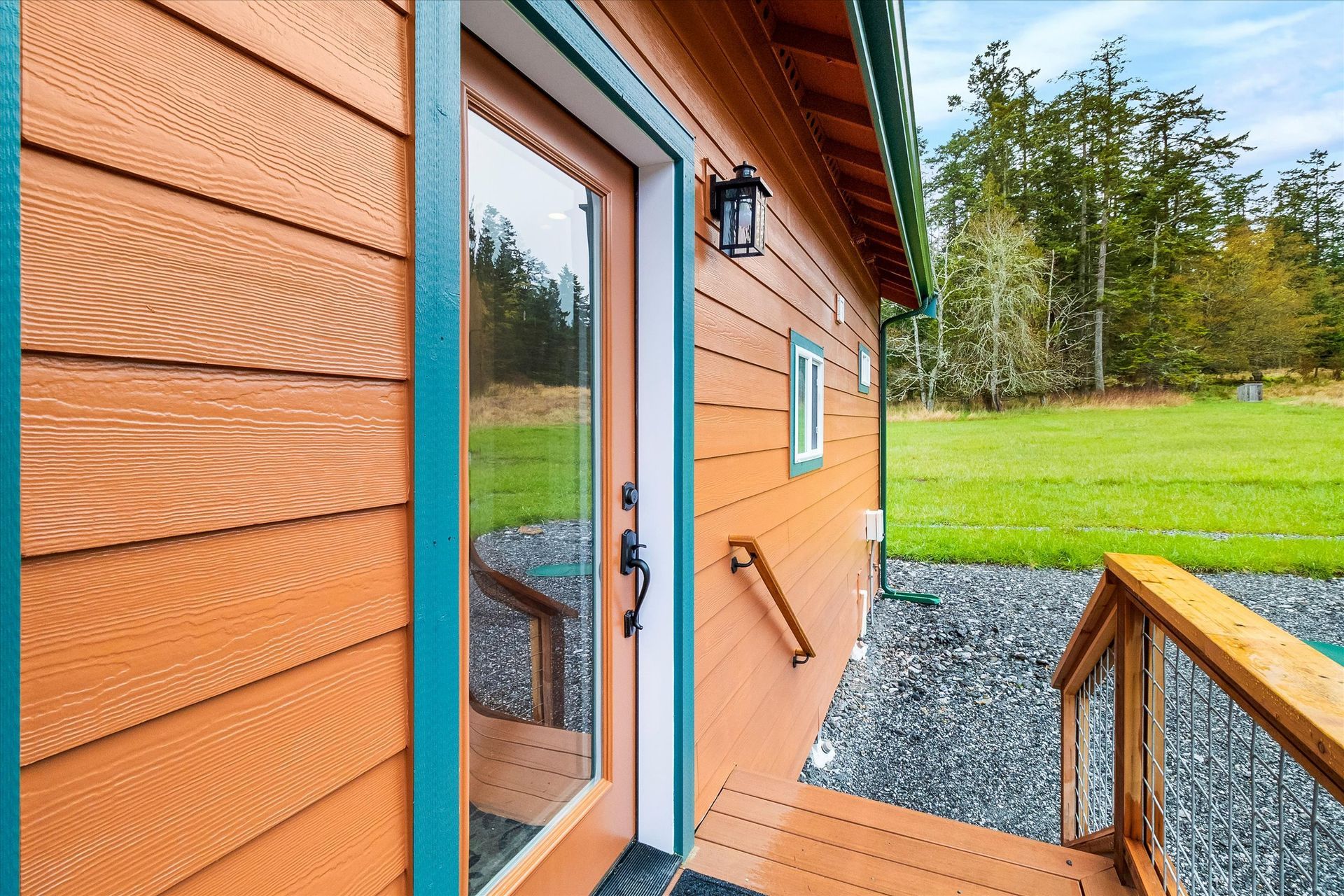 A side view of a cabin with orange horizontal siding, a green-trimmed glass door, a wooden deck, and a green field nearby.