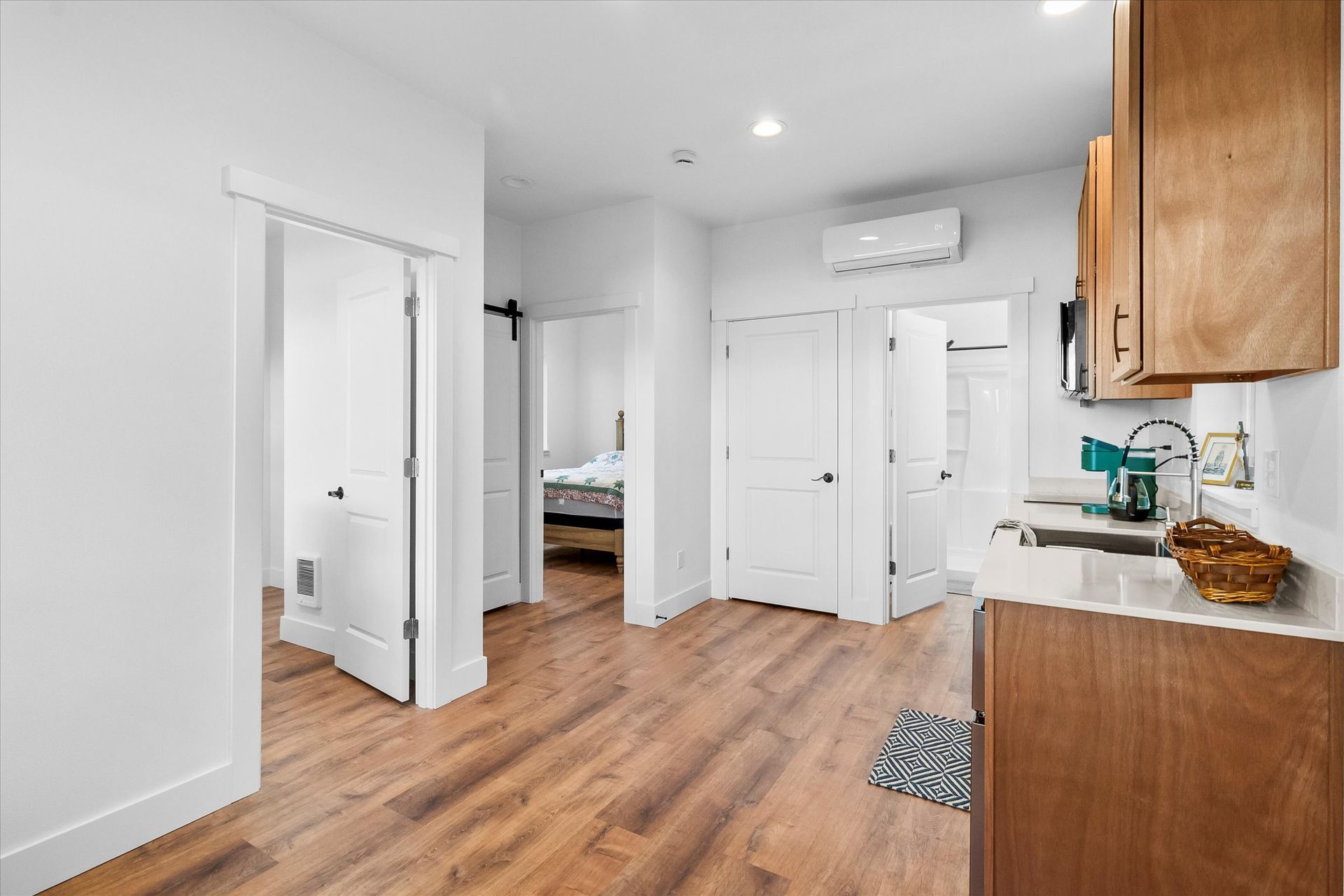 A bright interior view of an apartment, showing wood-look flooring, white doors, and a partial view of a kitchen area.