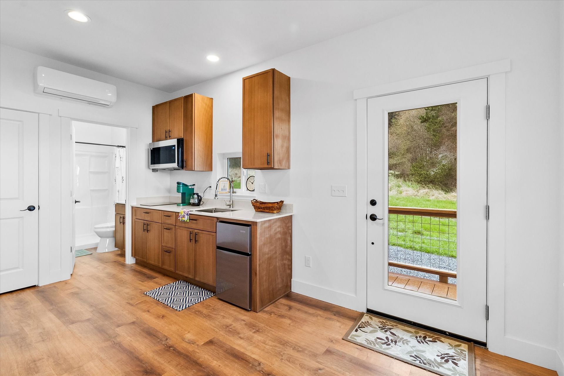 A small kitchen with wooden cabinets, a sink, microwave, and mini-fridge, leading to a bathroom and a glass-paneled door.