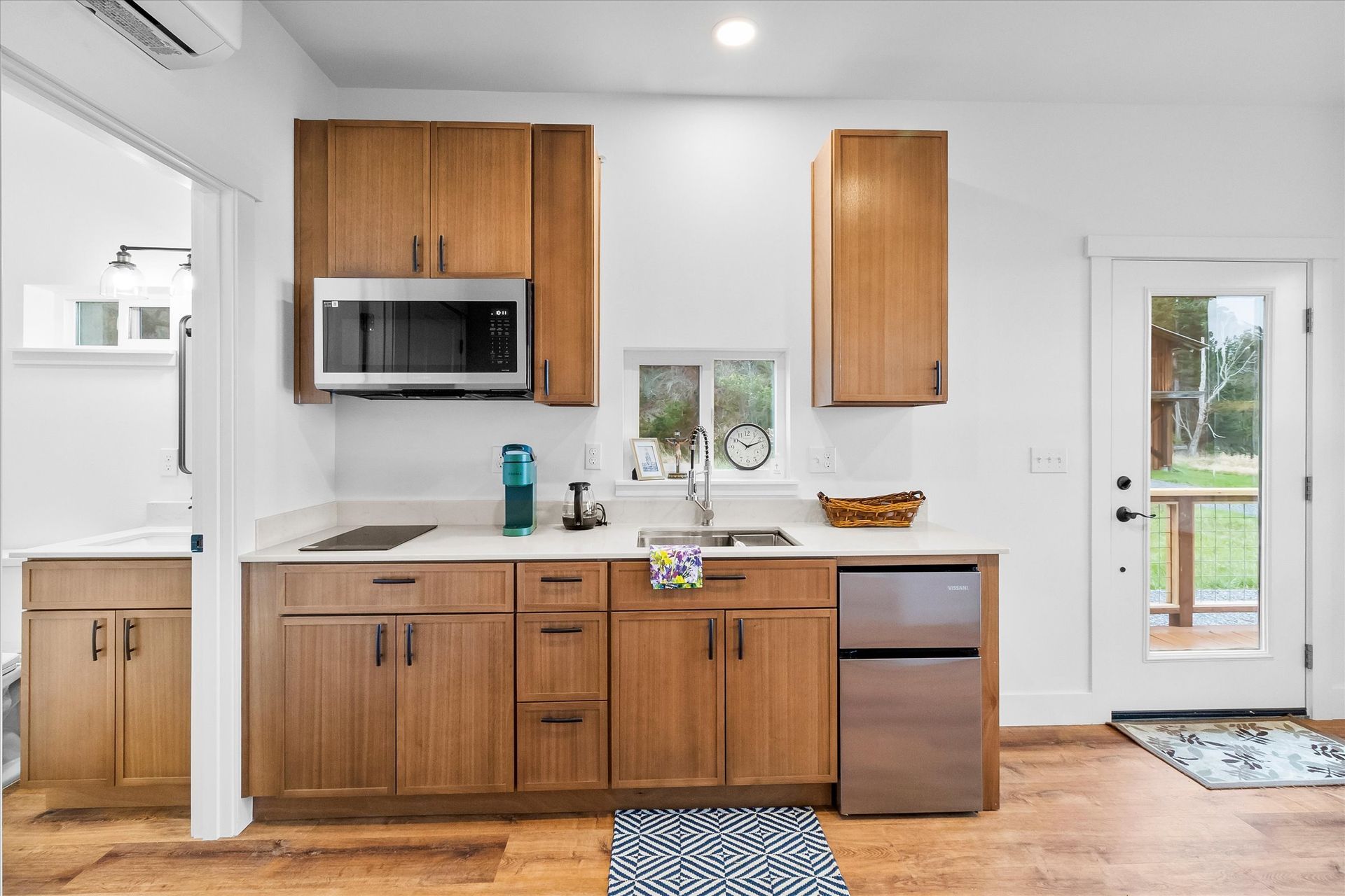 A small, modern kitchen with light wood cabinets, stainless steel appliances, and a white door leading outdoors.