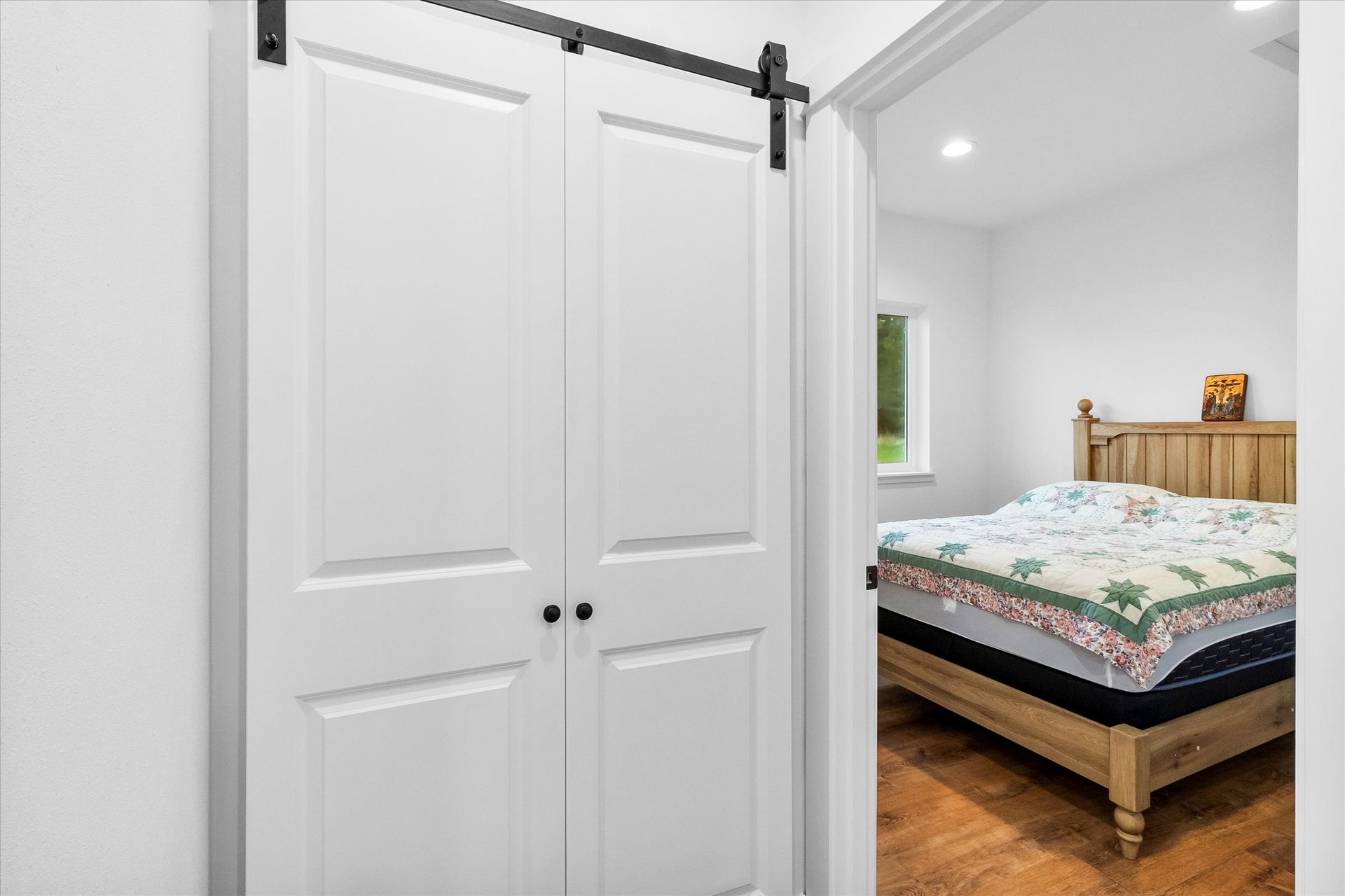 A white double barn door with black hardware sits next to a doorway looking into a bedroom with a wooden bed frame.