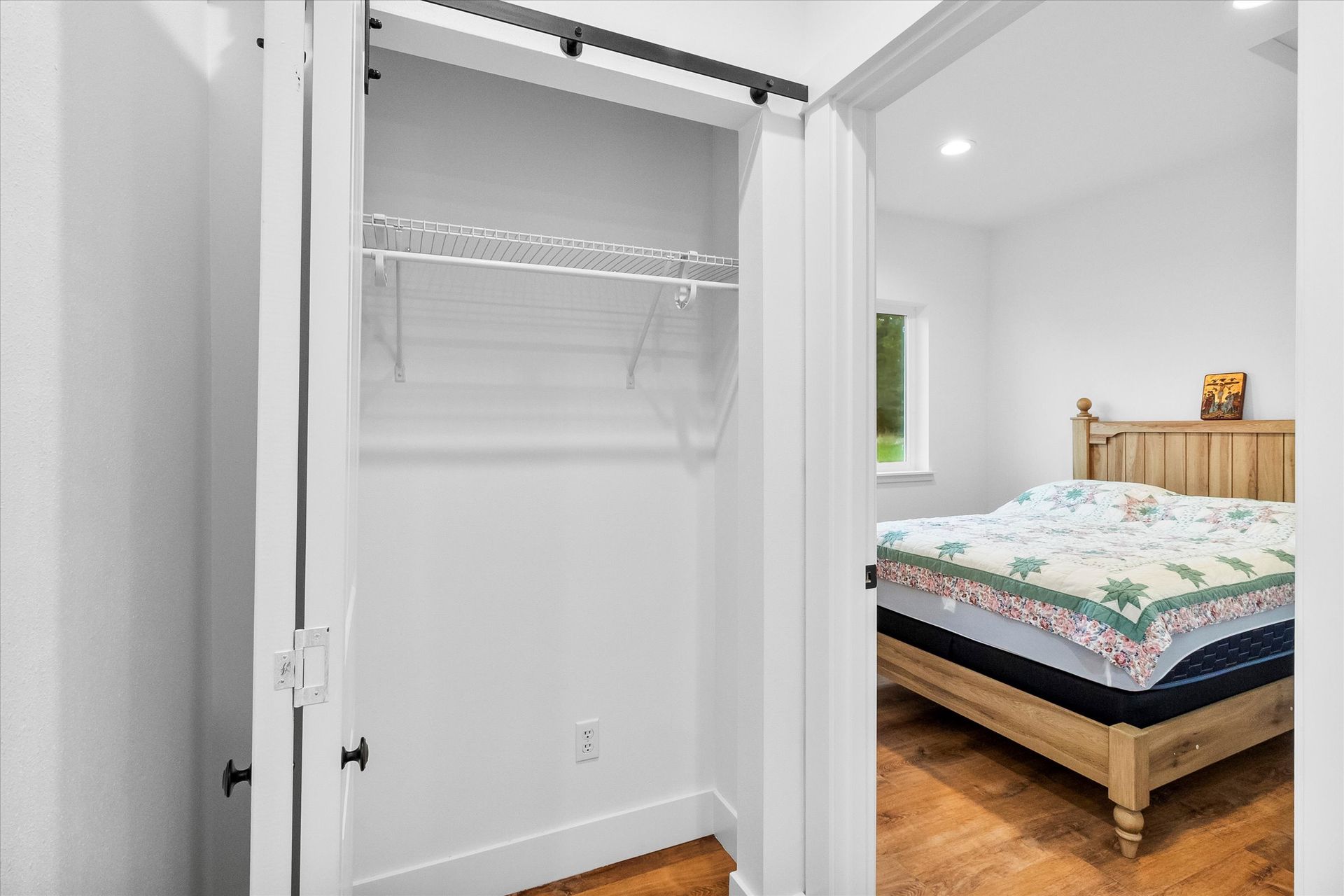 An open closet with a wire shelf sits next to a doorway showing a bedroom with a wooden bed frame and a patterned quilt.