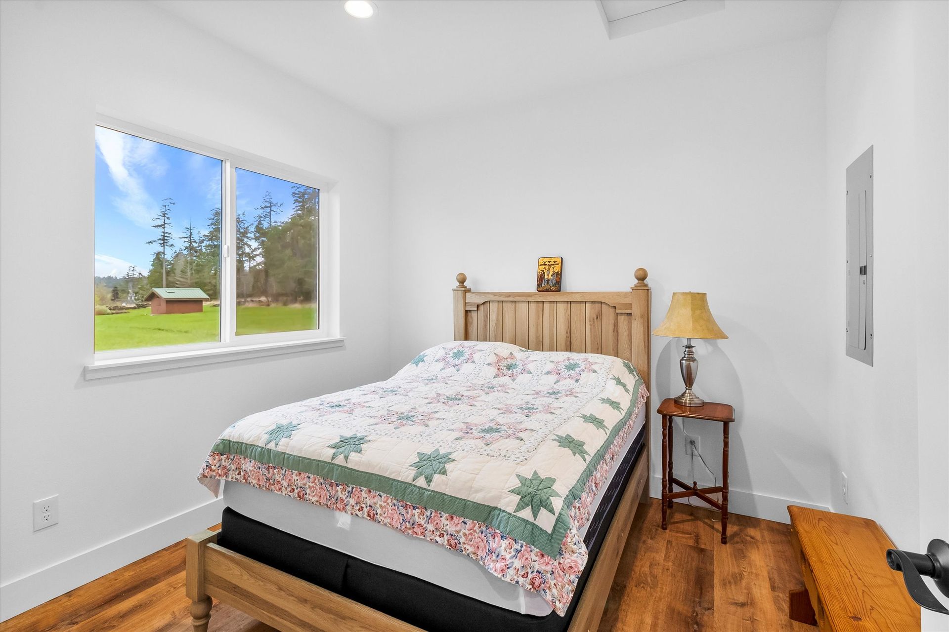 A simple bedroom with a wooden bed frame, a floral quilt, a side table with a lamp, and a window overlooking a green yard.