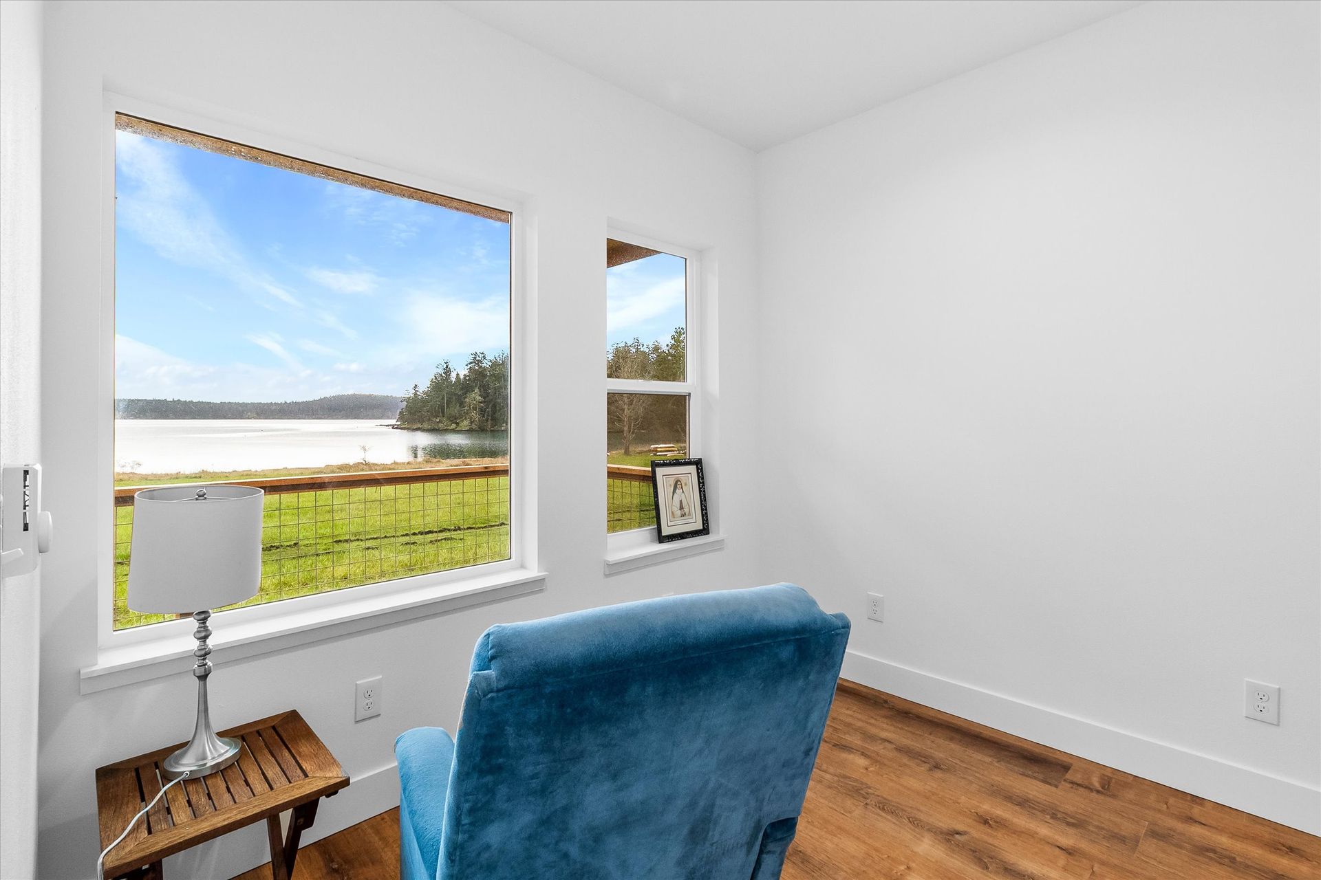 A blue armchair faces a large window overlooking a scenic lake and grassy yard in a minimalist room with white walls.