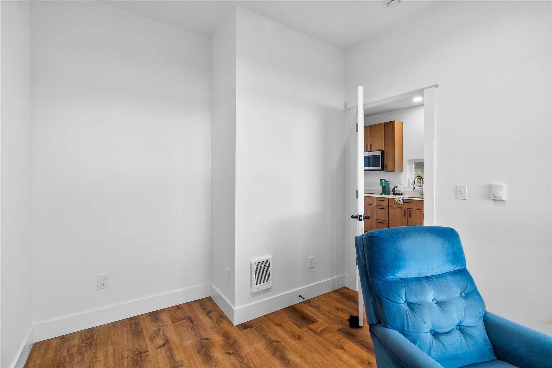 A blue velvet recliner sits in a corner of a bright, white room with wood flooring, facing an open doorway to a kitchen.