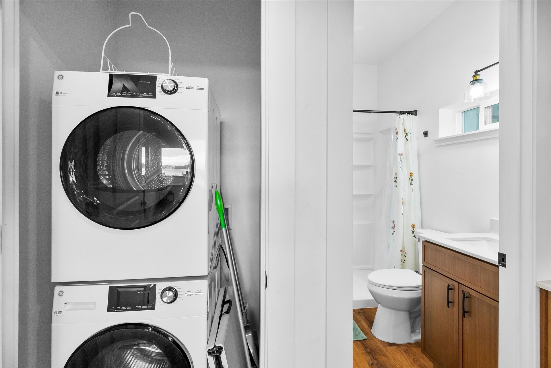 A closet with a stacked white washer and dryer next to a bathroom with a vanity, toilet, and shower curtain.