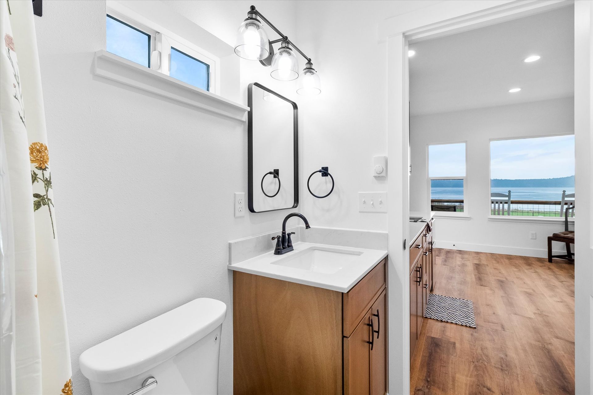 A small bathroom with a wooden vanity, mirror, and toilet, open to a room with wood floors and a scenic view of the water.