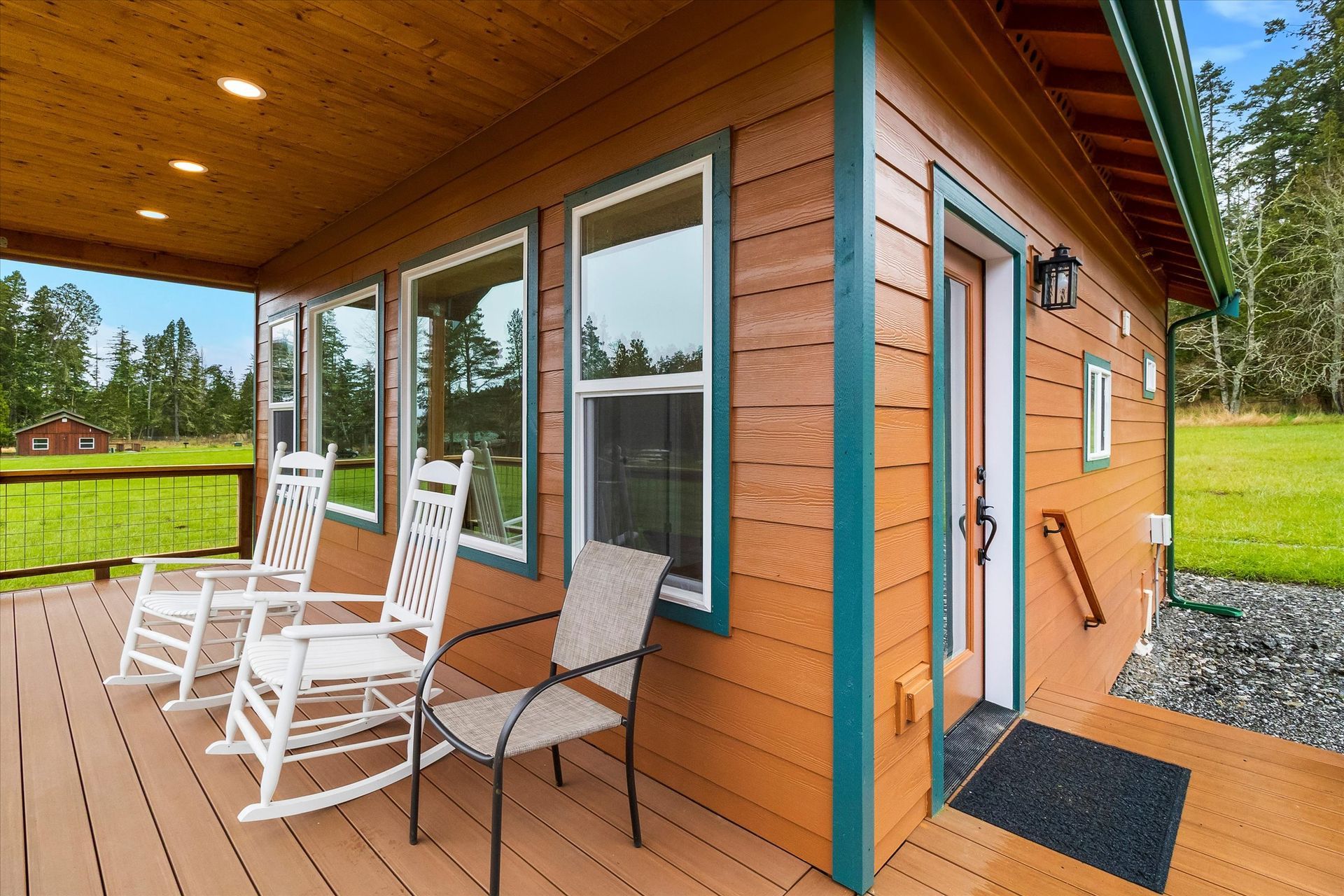 A wooden cabin deck featuring two white rocking chairs and a grey chair, set against a green field and forest backdrop.