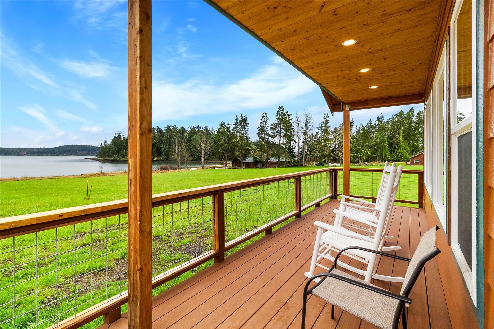 A wooden porch with two white rocking chairs overlooking a green field, a body of water, and trees under a blue sky.
