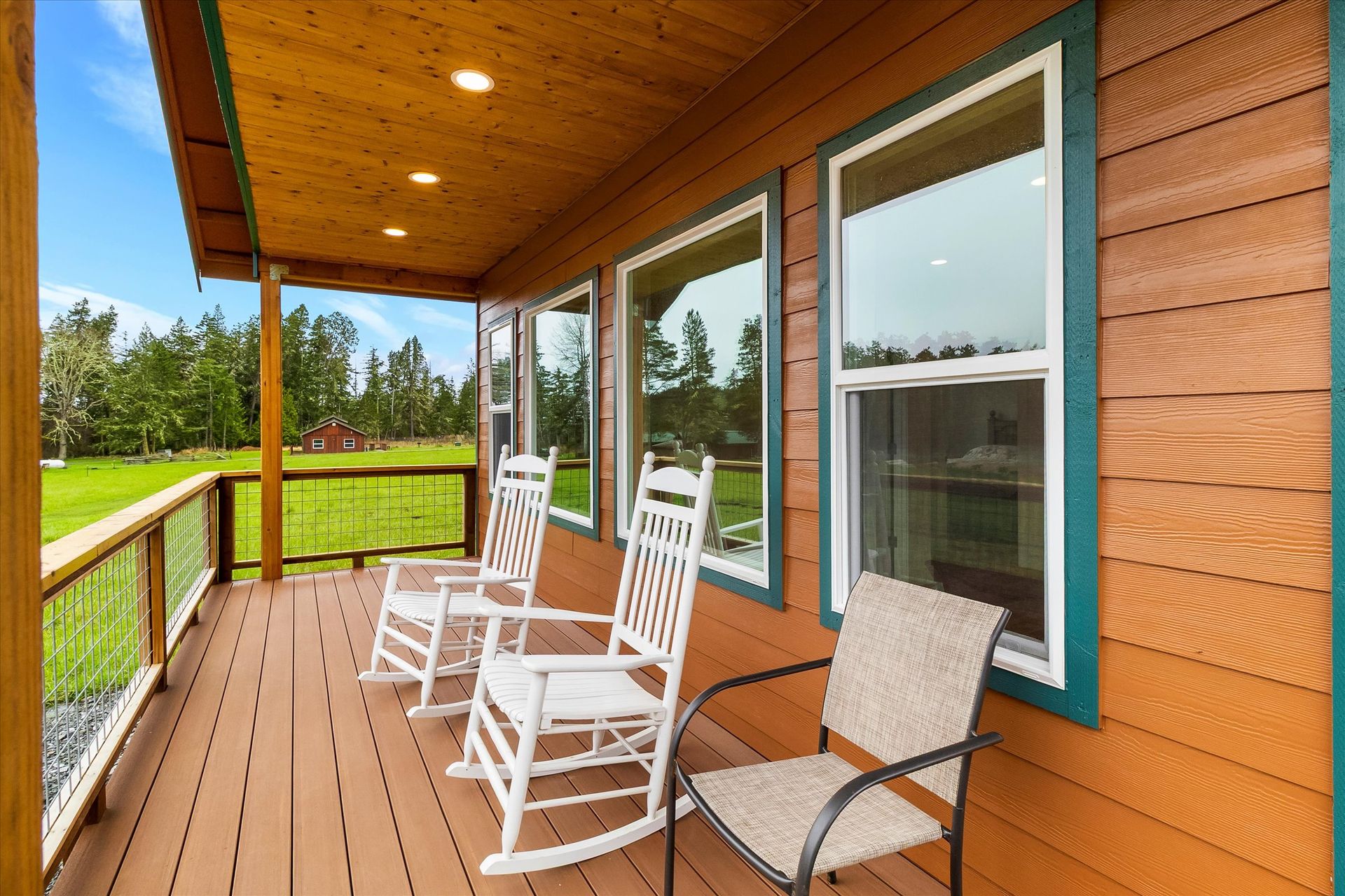 A covered wooden deck features two white rocking chairs and one neutral patio chair, overlooking a green grassy field.
