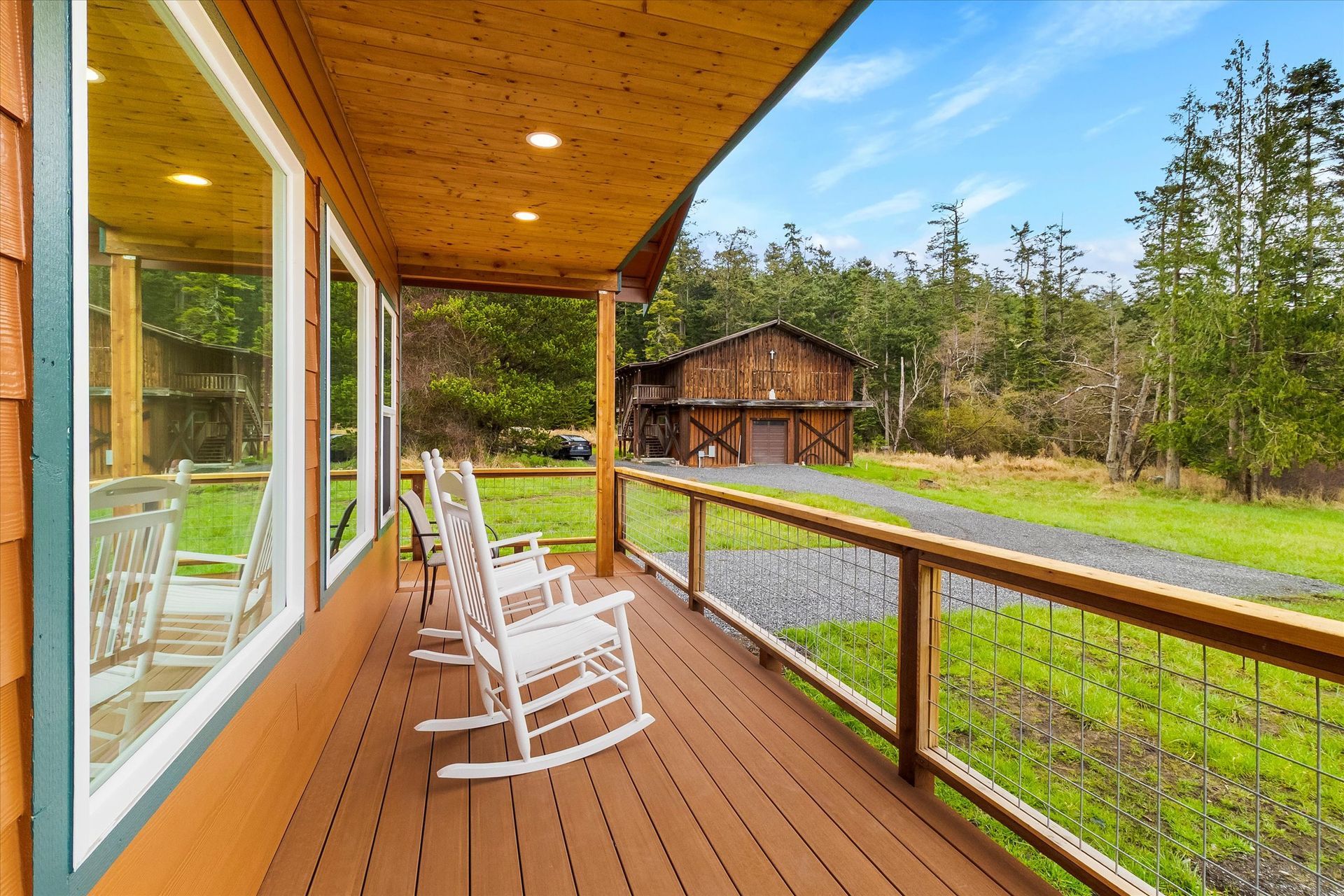 A covered wooden porch features three white rocking chairs, with a gravel path and a barn visible in the grassy landscape.