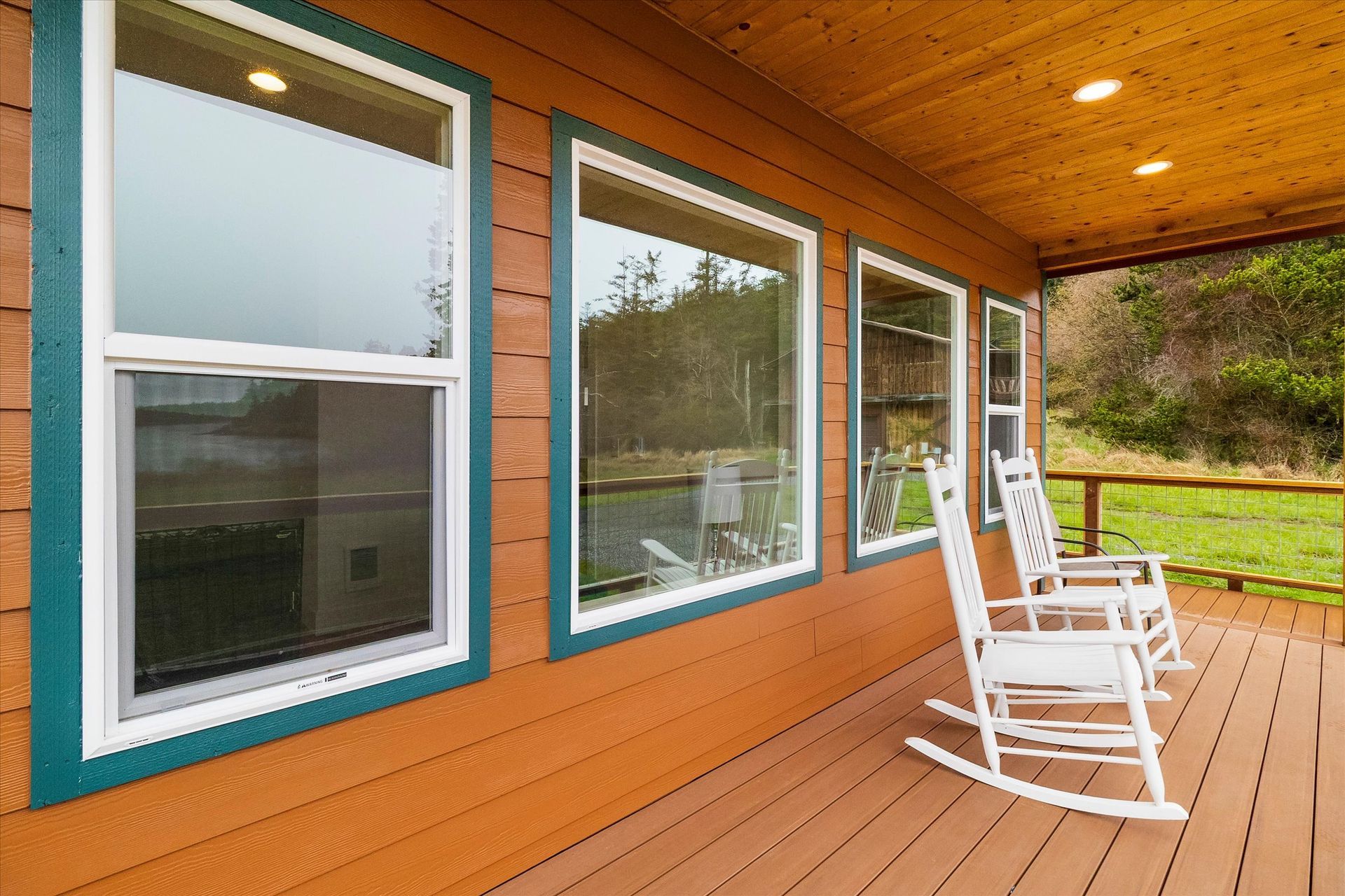 Two white rocking chairs on a wood deck outside a cabin with three large windows overlooking a grassy field.