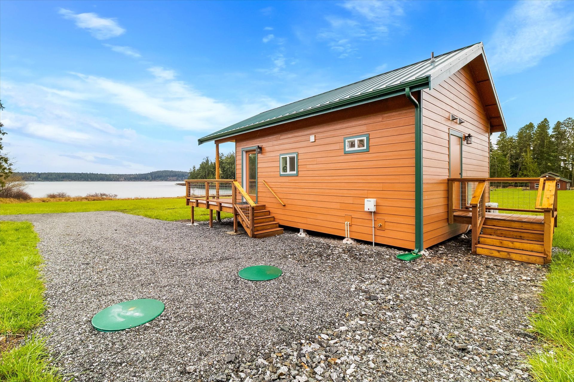 A wood-sided cabin with a green roof sits on a gravel lot overlooking a lake under a blue sky.