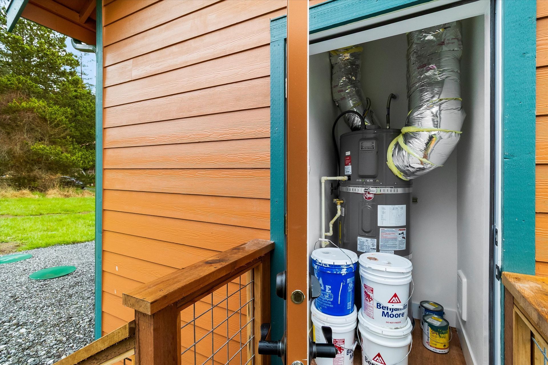 An outdoor closet on a wooden exterior house containing a water heater with attached ductwork and several storage buckets.