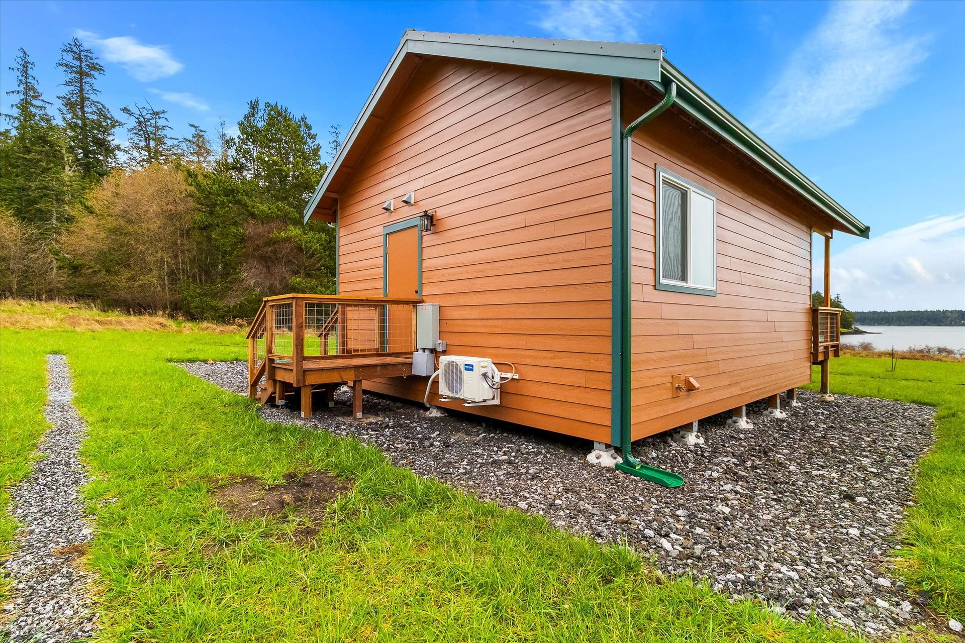 A small, wooden cabin with a green roof sits on a gravel pad near a grassy field and a body of water under a blue sky.