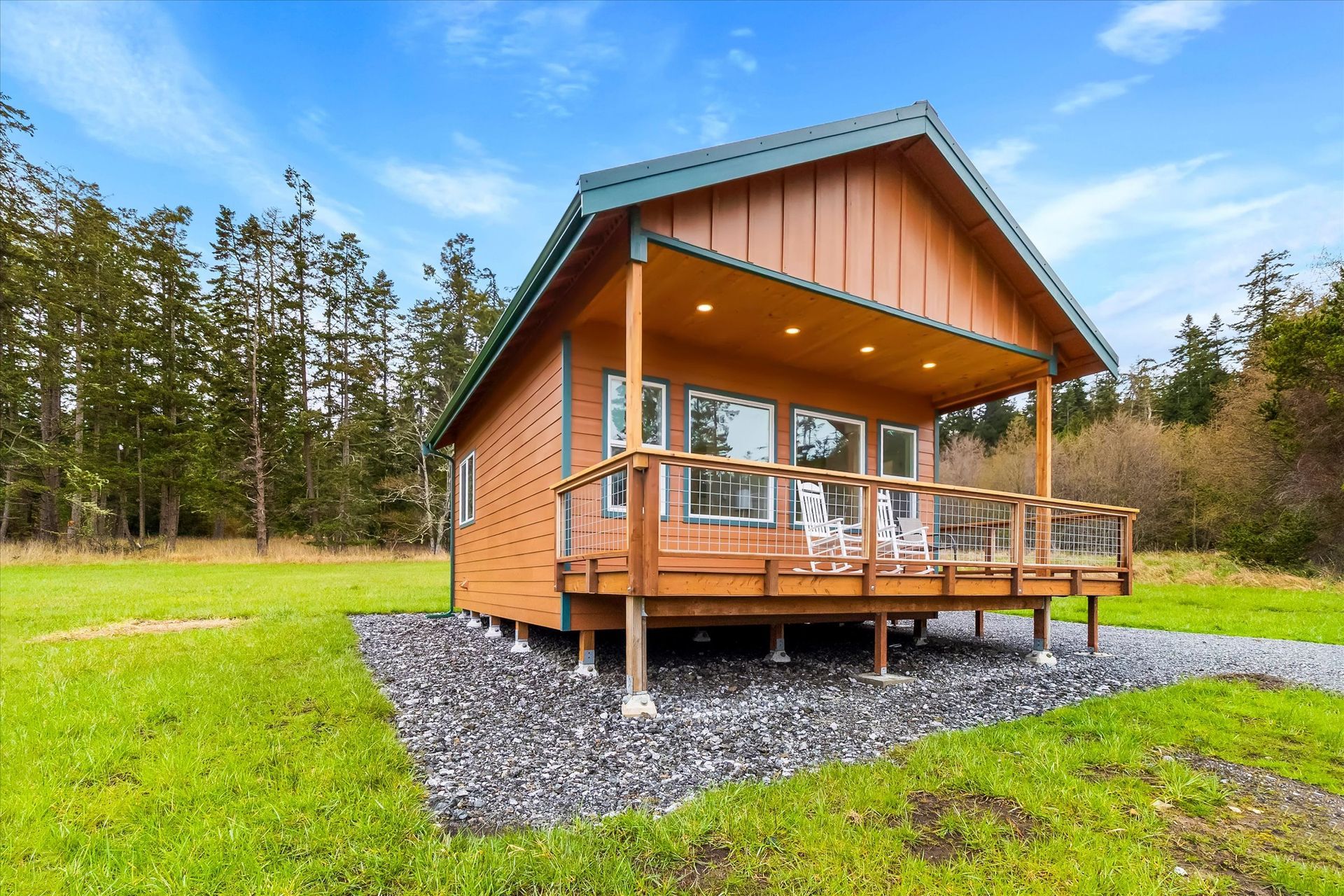 A small, brown wooden cabin with a covered deck and porch lights, situated in a field near a forest under a blue sky.
