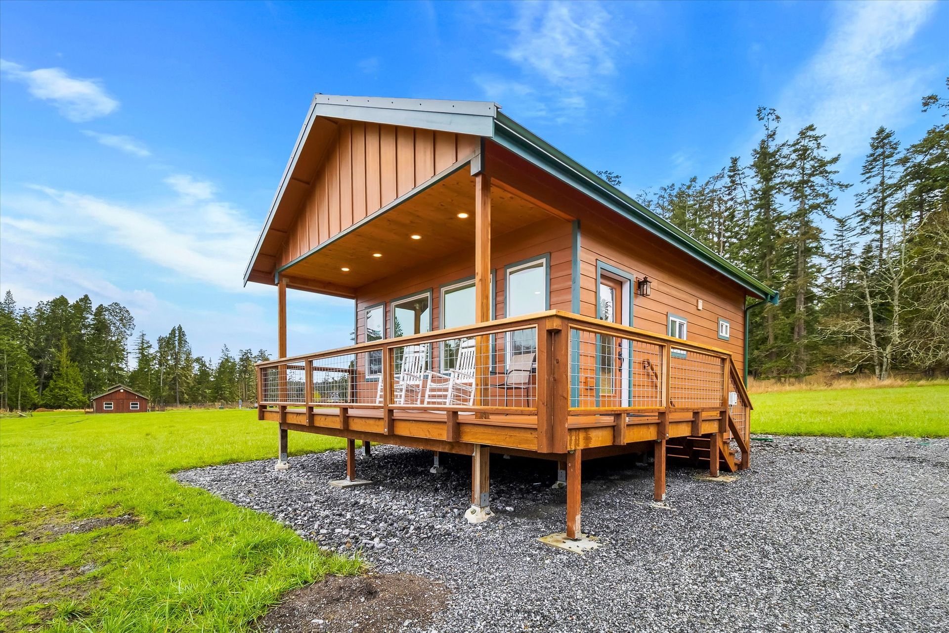 A rustic wooden cabin with a spacious deck, set on a gravel base surrounded by a green field and trees under a blue sky.