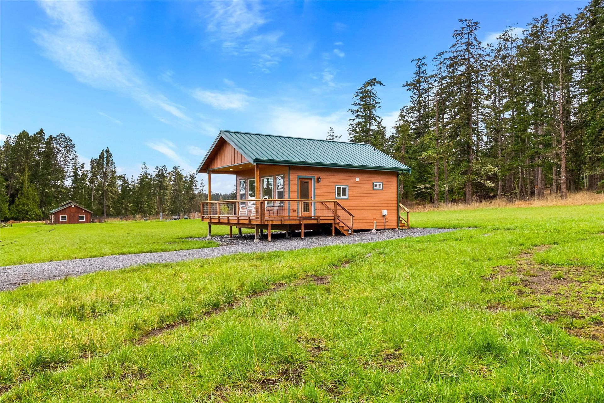 A small, brown wooden cabin with a green metal roof sits on a gravel path in a large, grassy field surrounded by trees.