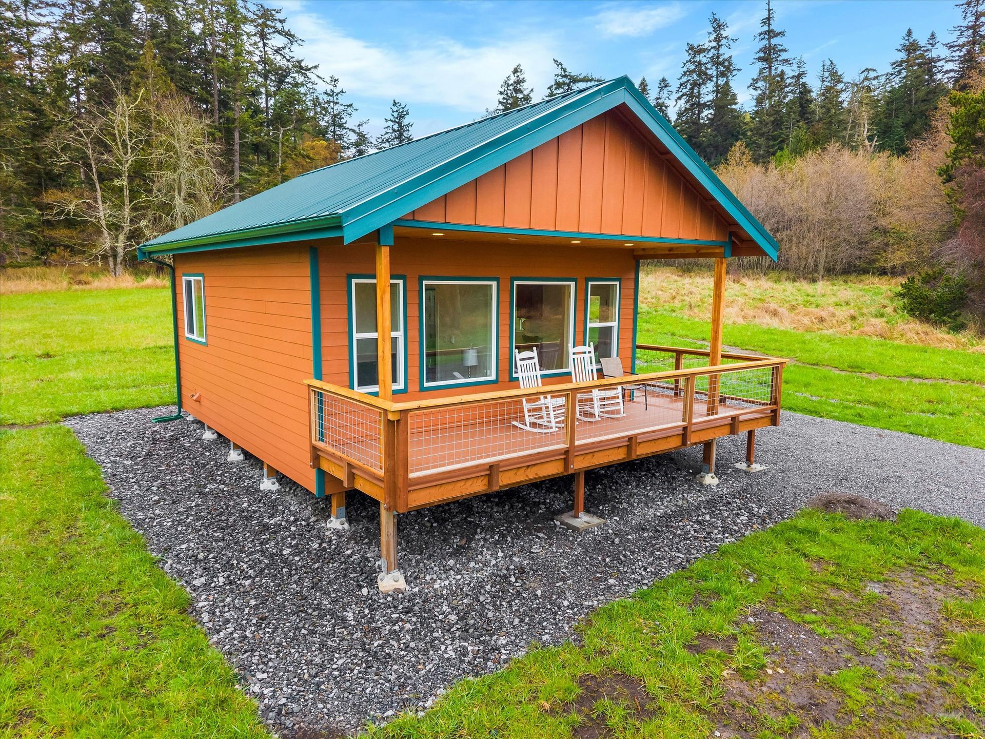 A small, orange cabin with a green metal roof and a wooden deck featuring two rocking chairs, set in a grassy field.