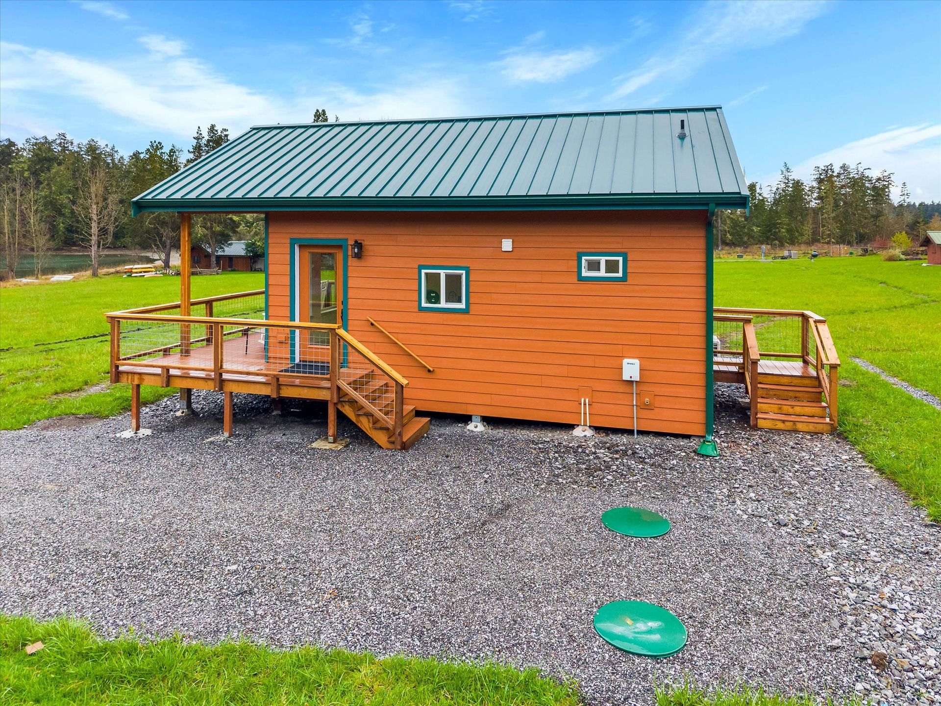 A small, orange cabin with a green metal roof, a wooden deck, and two green circular septic lids on a gravel lot.