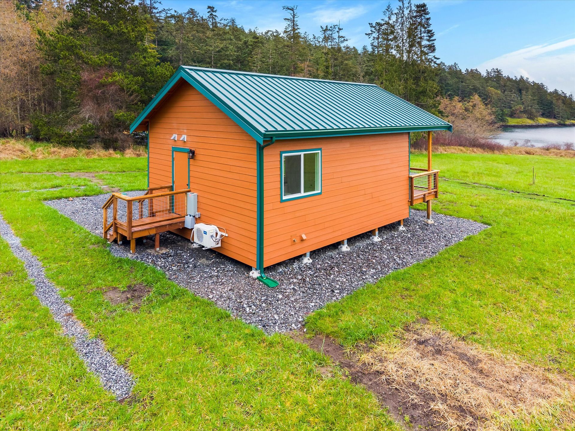 An orange, single-room cabin with a green metal roof and small wooden porch sits on a gravel pad in a grassy, rural field.