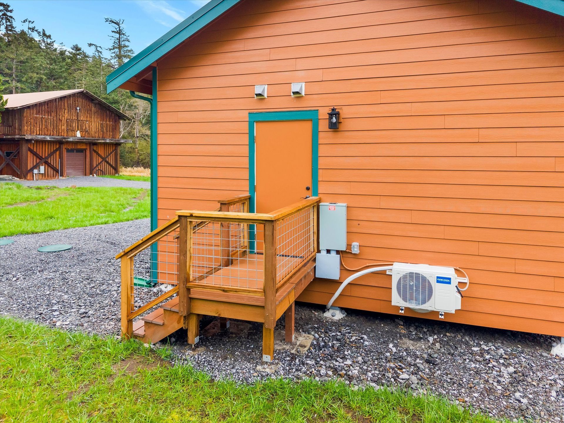 A side view of a bright orange building with a wooden deck and staircase, near a rustic barn on a gravel lot.