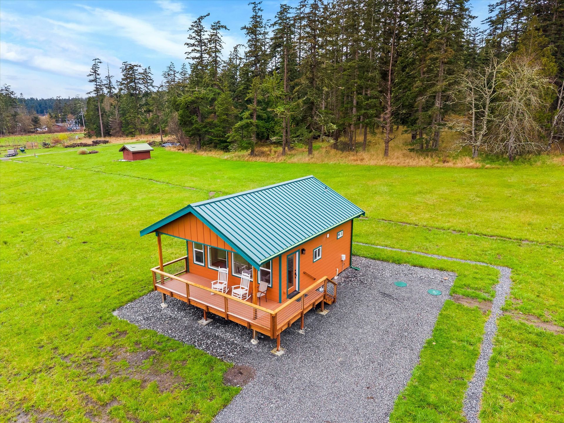 An elevated orange cabin with a green metal roof and front porch sits on a gravel pad in a large, grassy field near trees.