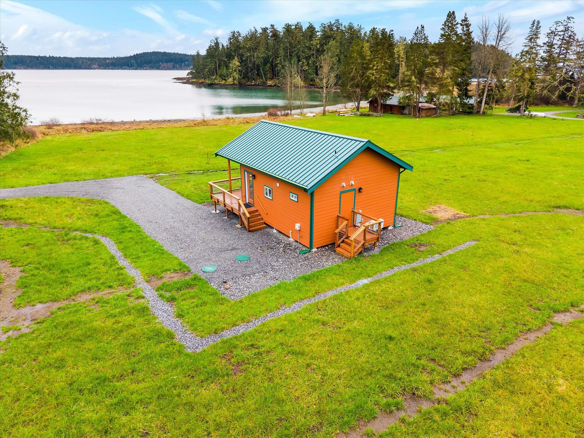 A small orange cabin with a green metal roof sits on a gravel pad in a large, grassy field overlooking a calm bay.