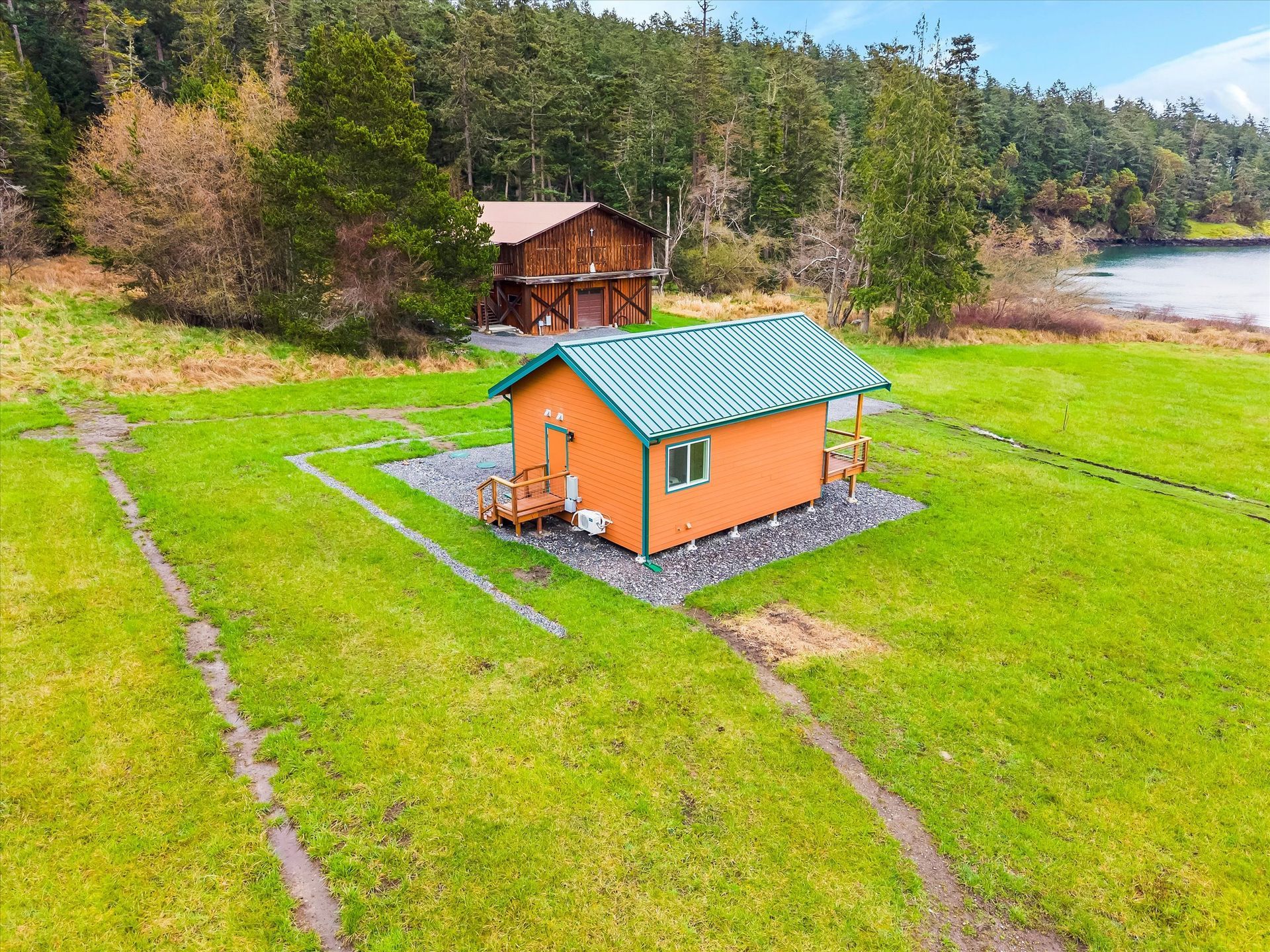 An orange cabin with a green metal roof sits on a gravel patch in a grassy field, with a large wooden barn in the back.