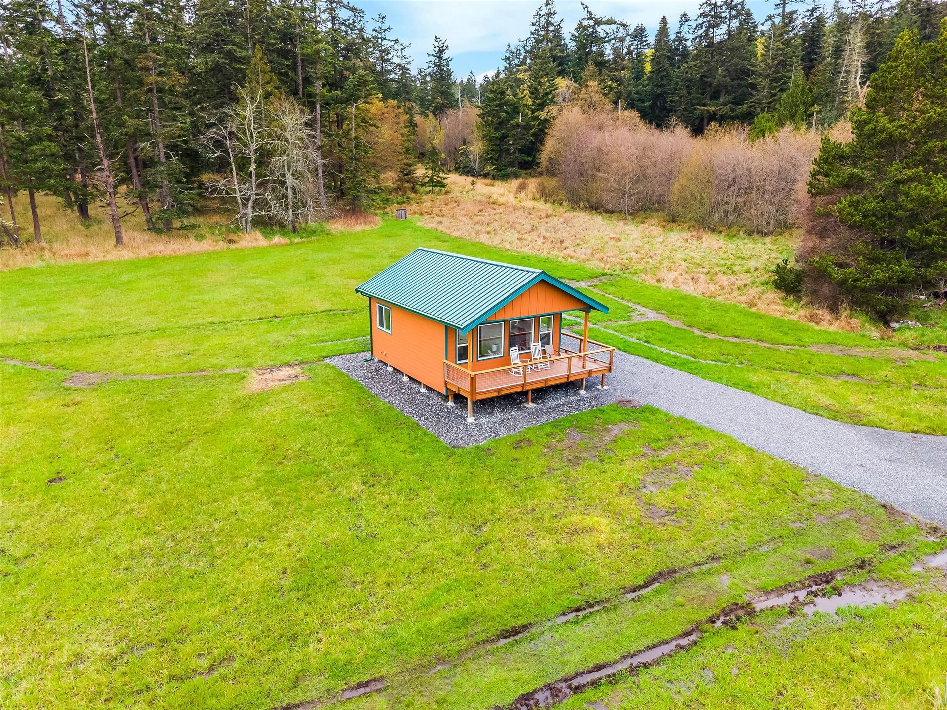 An aerial view of a small orange cabin with a green corrugated metal roof, situated in a large grassy field near woods.