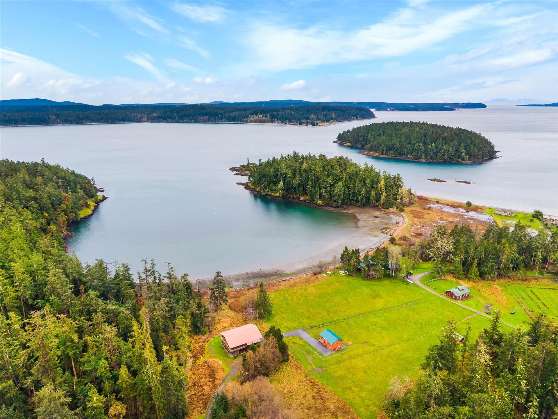 An aerial view of a coastal landscape featuring a green meadow with two small houses beside a bay, trees, and small islands.