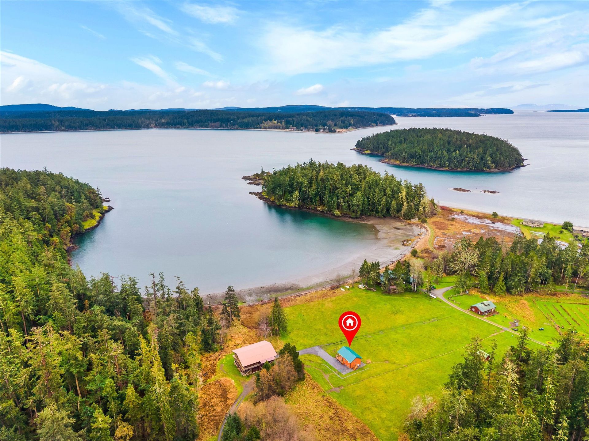 An aerial view of a house with a red map pin on a grassy property overlooking a bay with forested islands.
