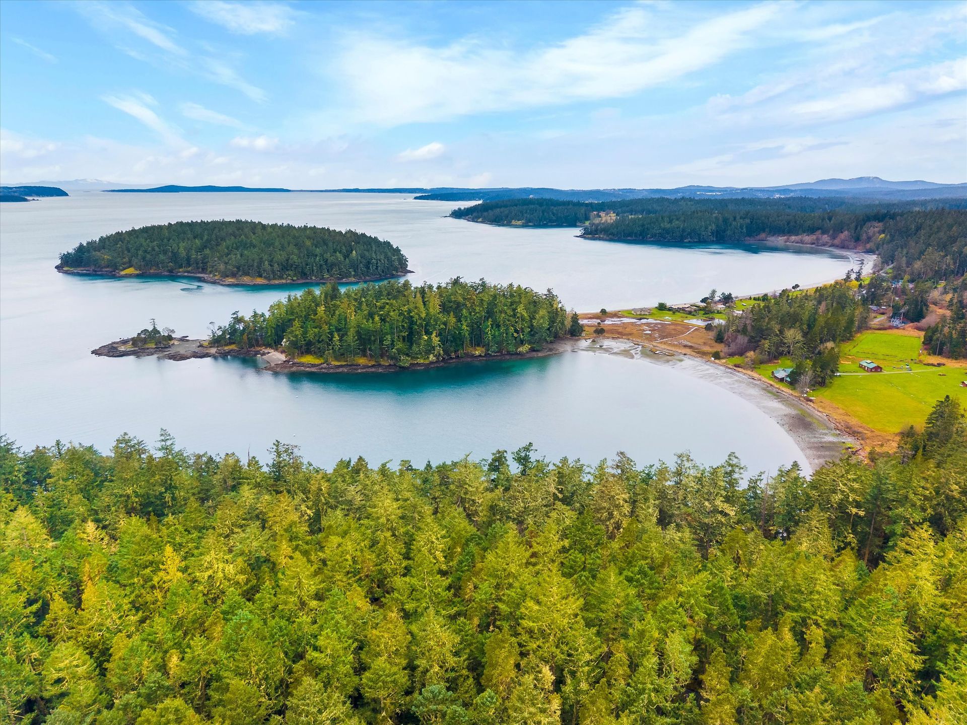 An aerial view of a forested coastline with several small, tree-covered islands in a calm, blue bay under a bright sky.
