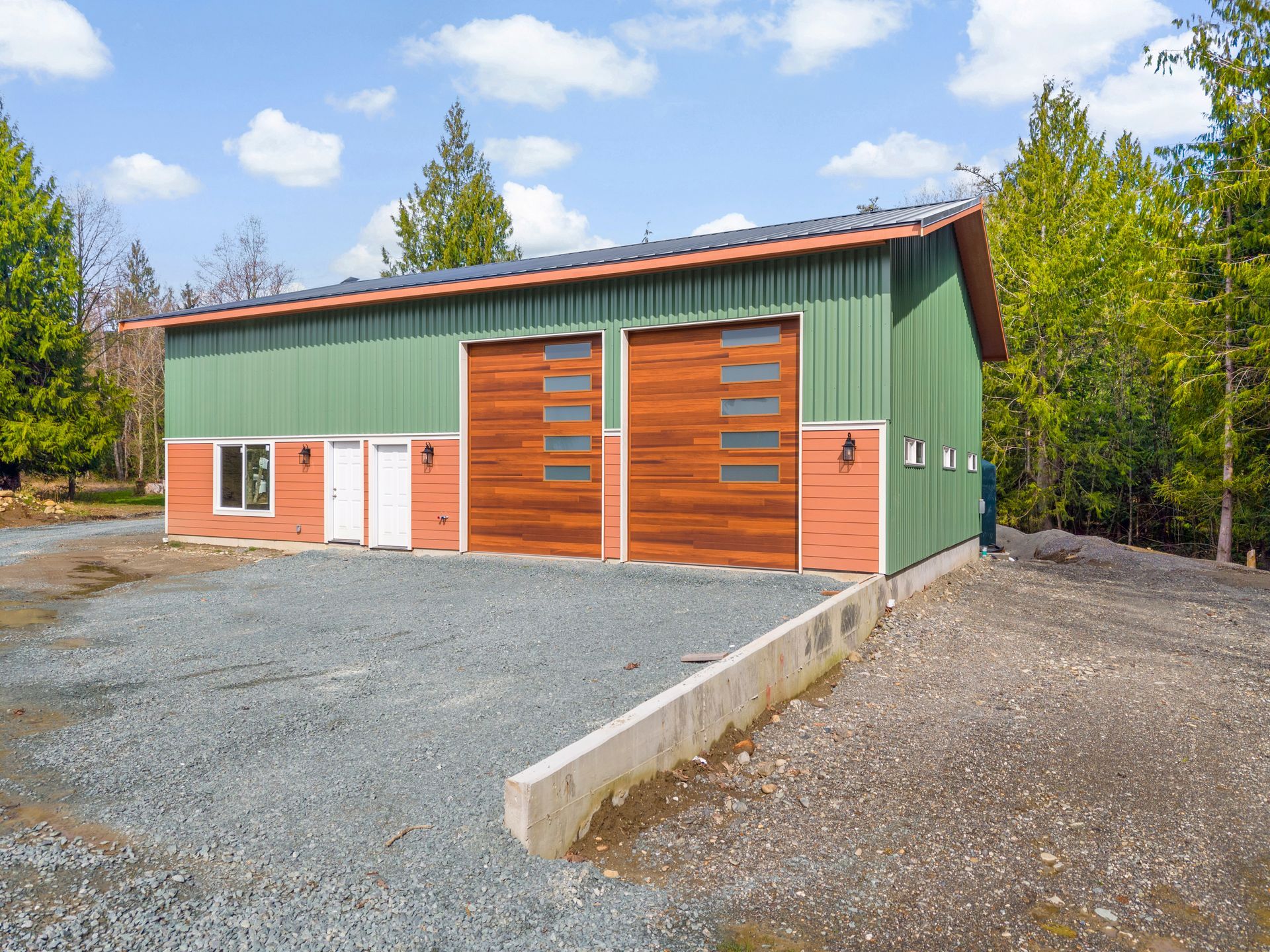 A green, two-story metal workshop building with two wooden garage doors, white doors, and windows, set in a wooded area.