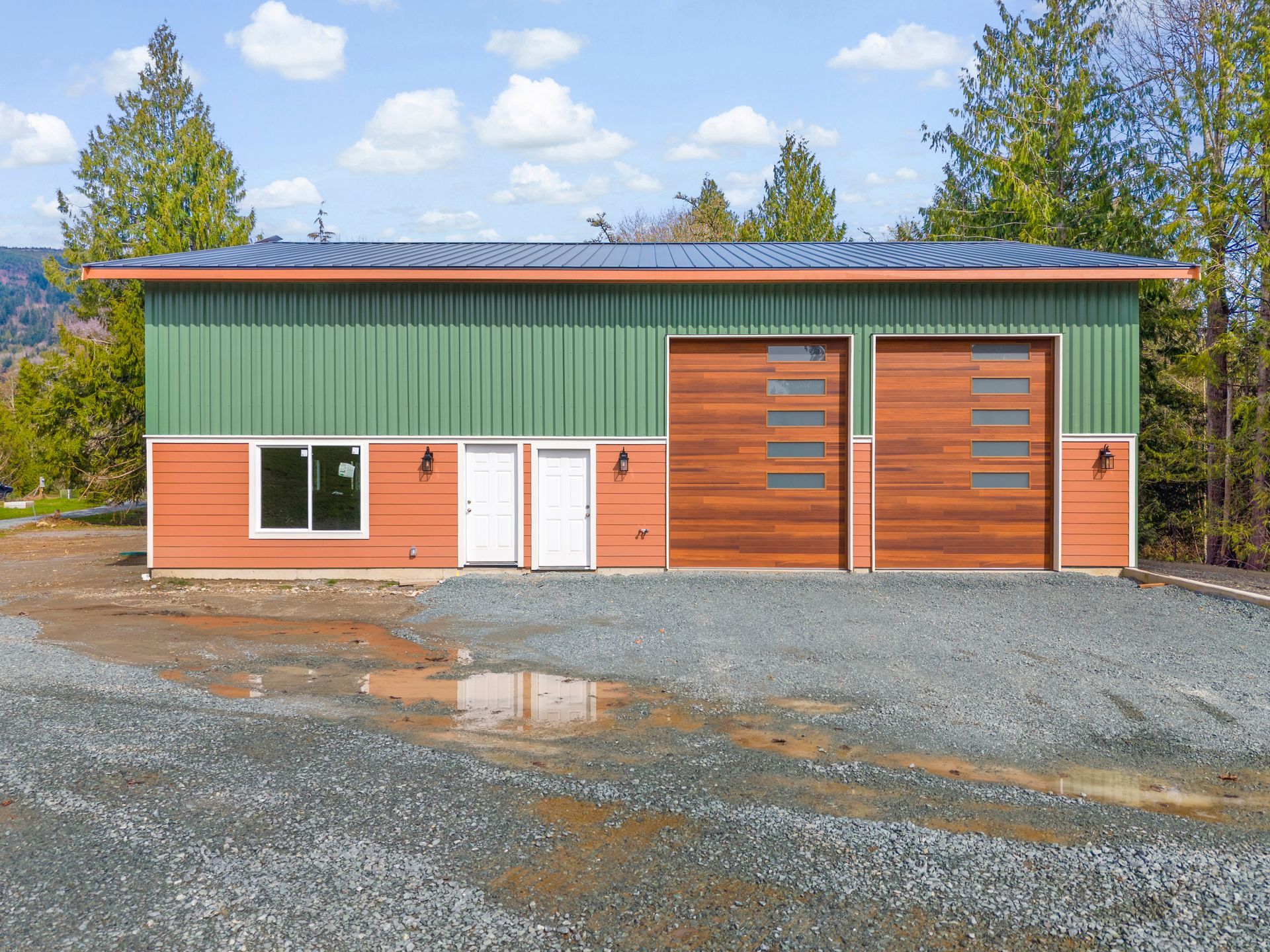 A modern, rectangular workshop building with green metal siding above orange horizontal panels, and two wooden garage doors.