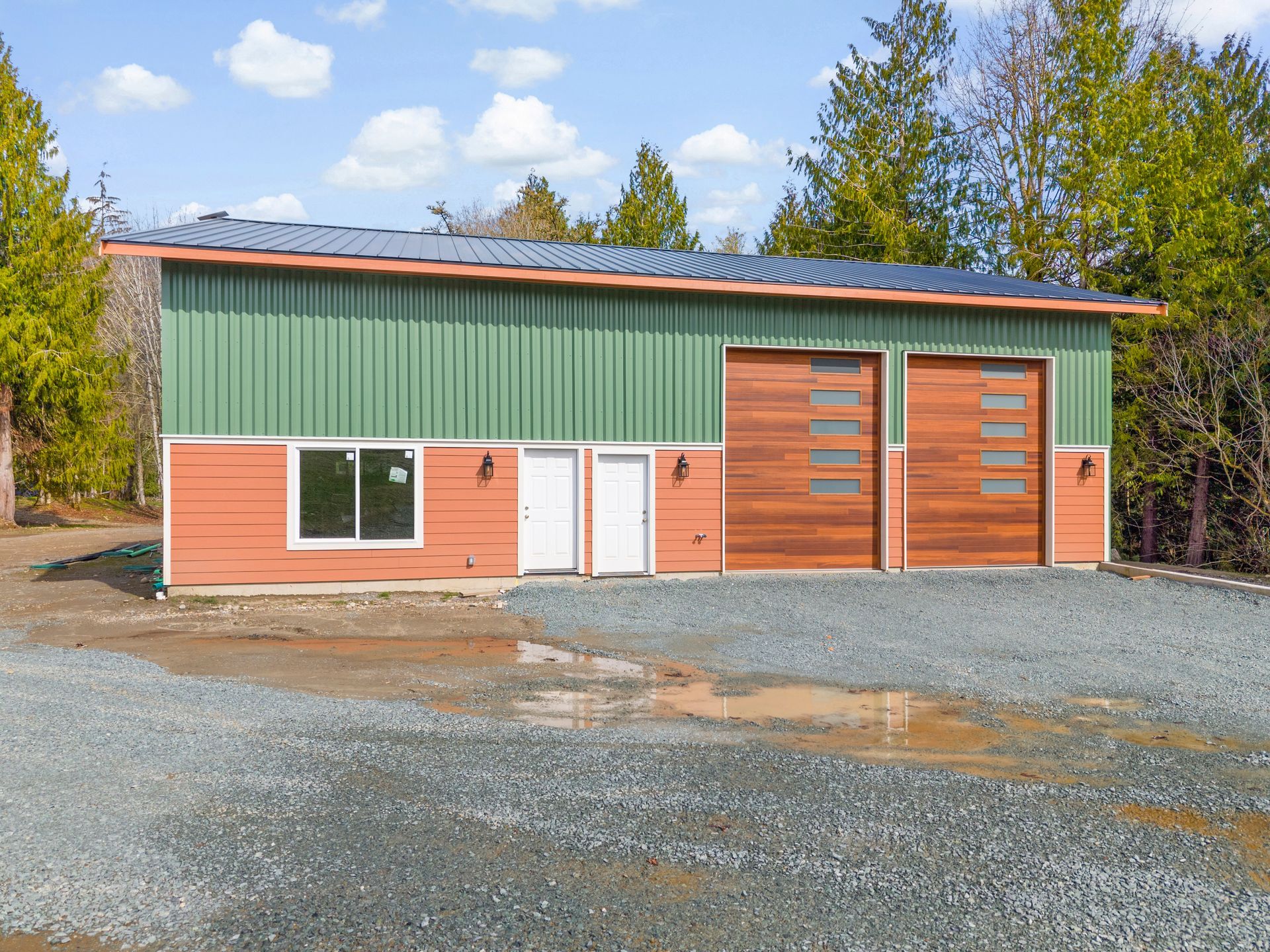 A single-story metal building with green siding above orange horizontal panels, two garage doors, and a window.