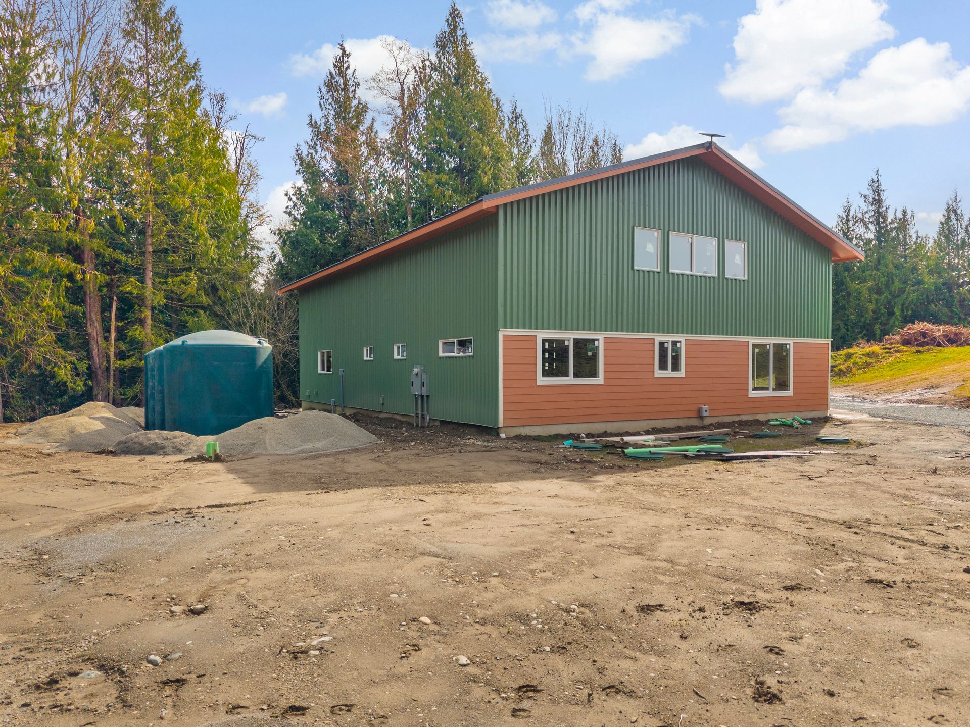 A green metal building with a tan lower exterior, surrounded by forest and a green water tank on a dirt lot.