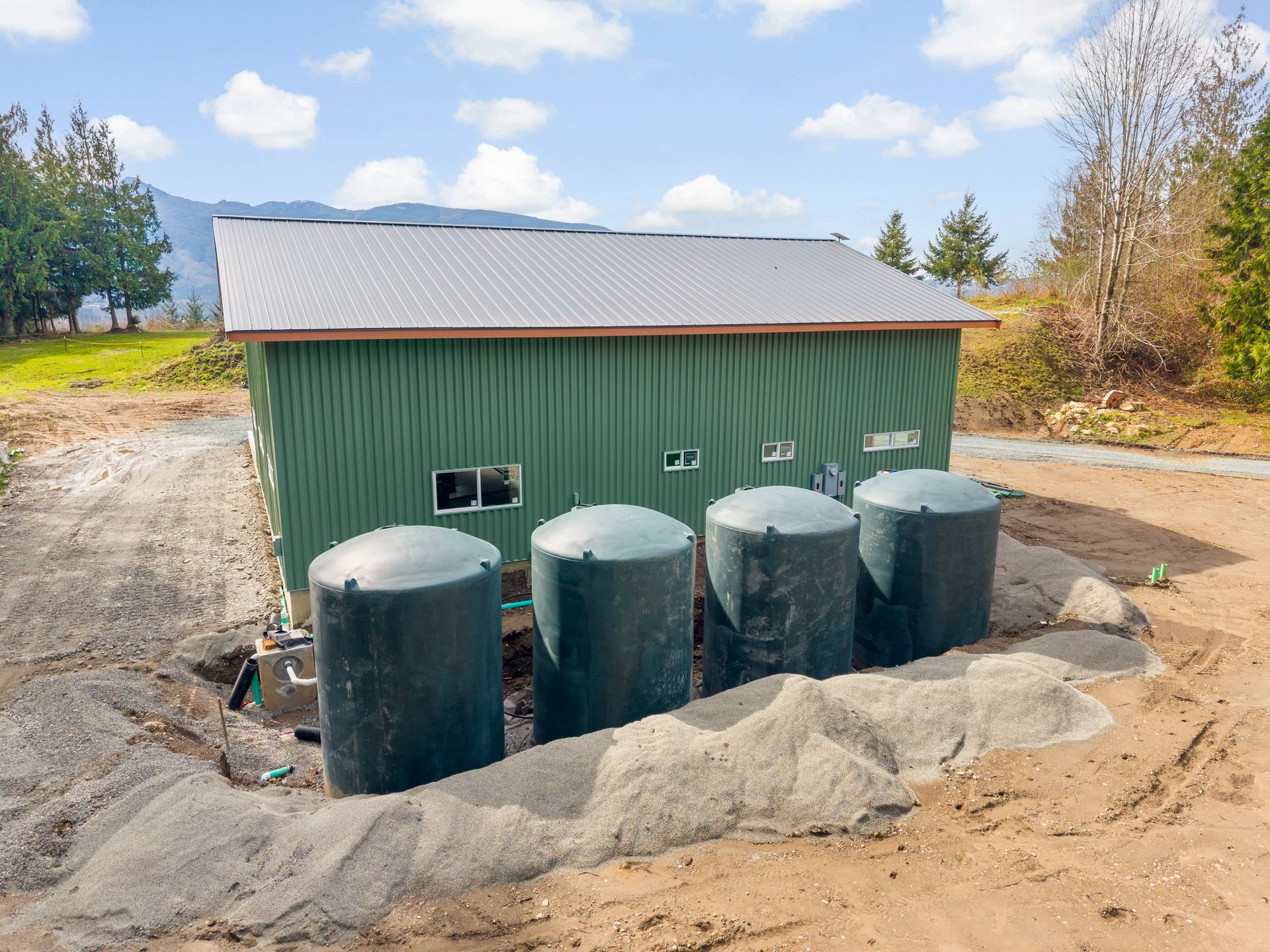 A green metal building with four dark green vertical water tanks sitting in a sandy, graveled area with trees nearby.